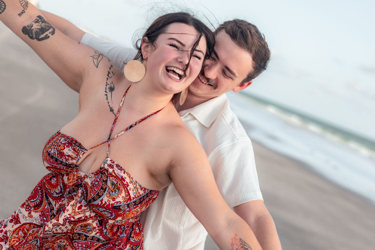 Laughing couple in a red floral dress and white shirt embrace with arms outstretched on a sandy beach at sunset.