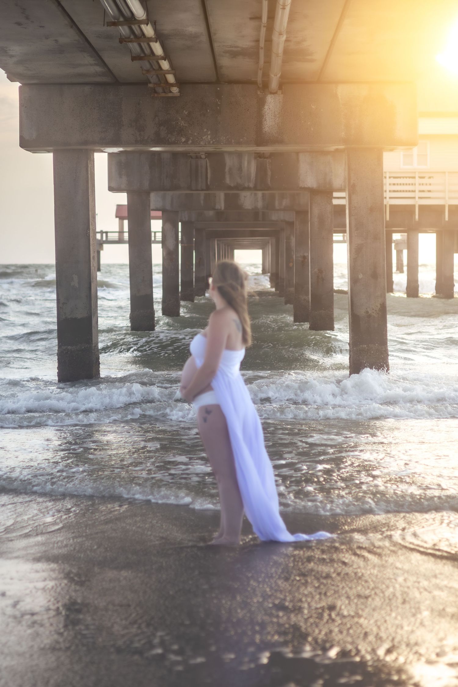 Expecting mother in a flowing white gown stands barefoot in the surf beneath a beach pier at sunrise.