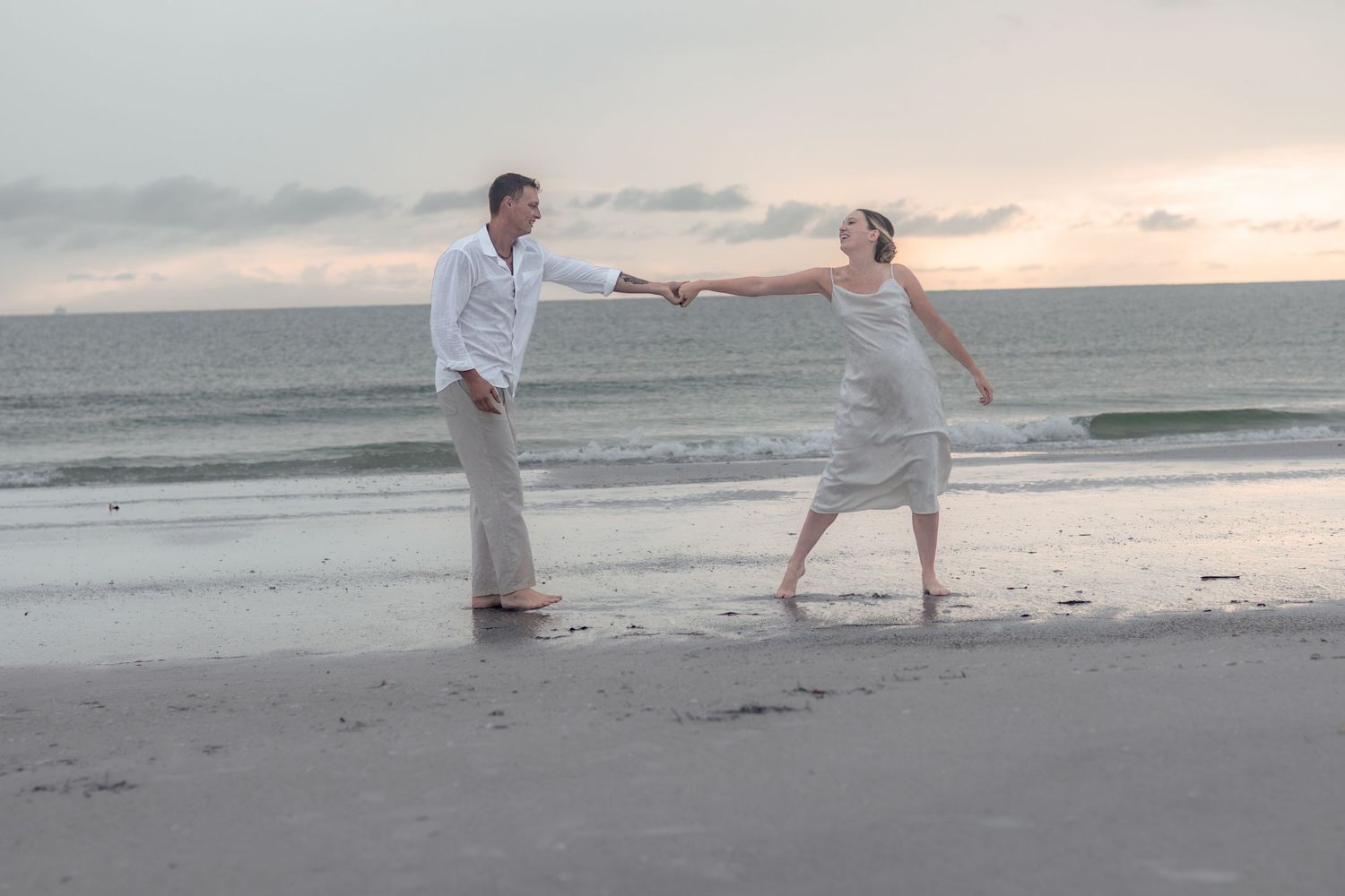 Couple holds hands at arm's length while playfully walking barefoot along the shoreline under a pastel cloudy sky.