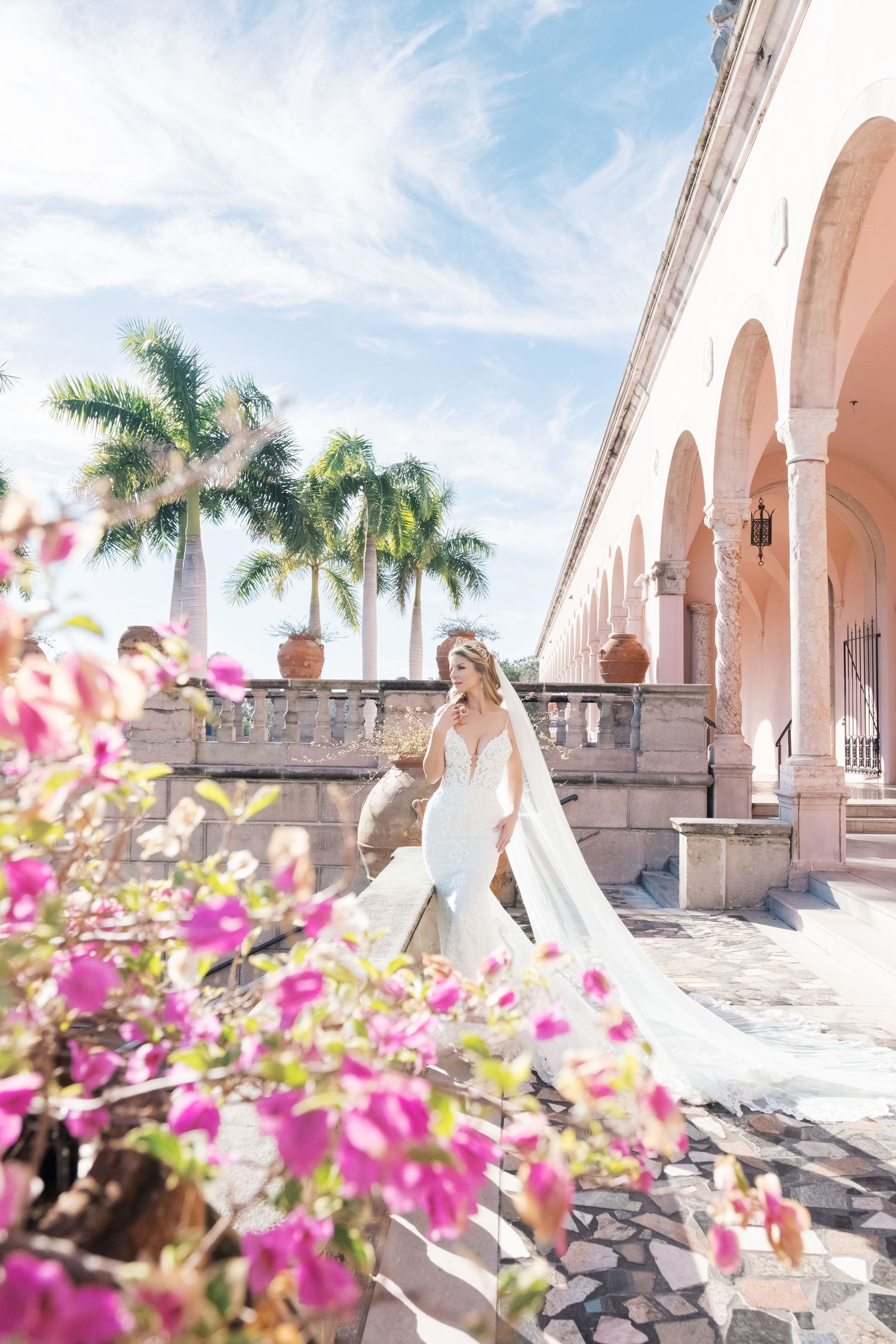Bride in white wedding dress poses gracefully under Mediterranean-style arches with palm trees and pink flowers. UK wedding photographer