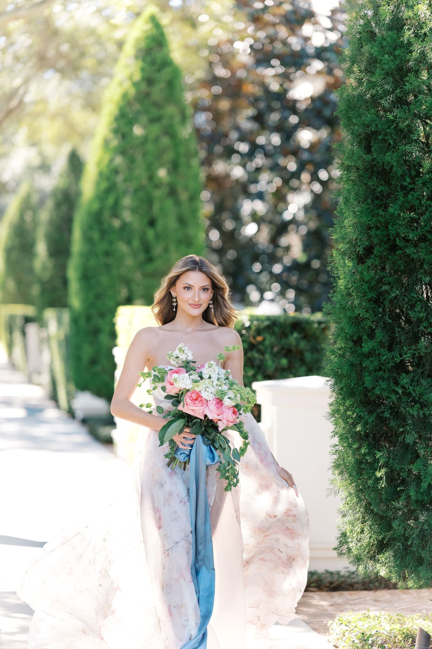 A dreamy bridal portrait in a garden with tall cypress trees, featuring a flowing blush gown and pink bouquet. Wedding uk photographer.