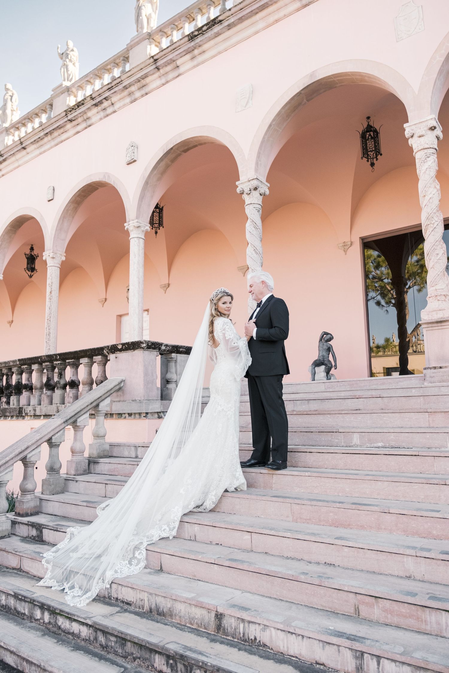 A couple in wedding attire poses on grand pink stone steps with white archways and columns in an elegant outdoor setting. London uk session.