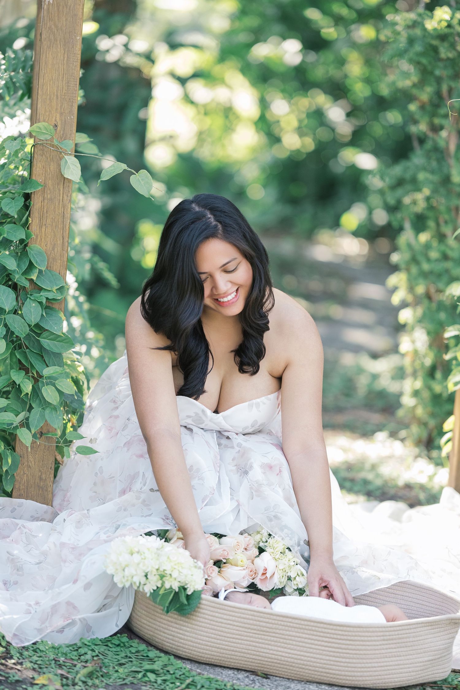 A woman in a white dress holding newborn surrounded by lush greenery in a dreamy outdoor setting. motherhood photographer, london uk