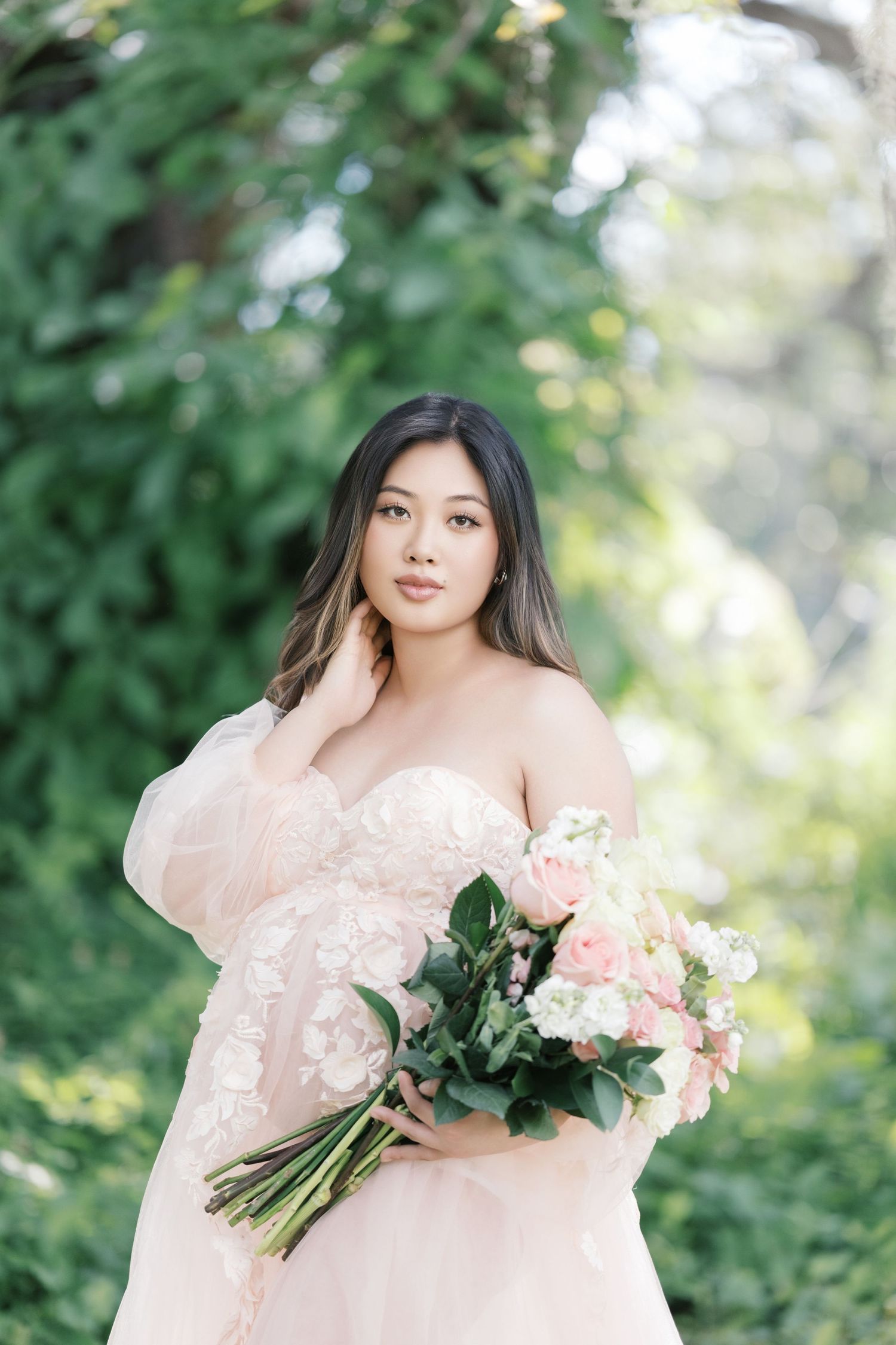 A maternity session in a soft pink off-shoulder dress poses outdoors holding a bouquet of pink and white flowers in a garden setting. UK