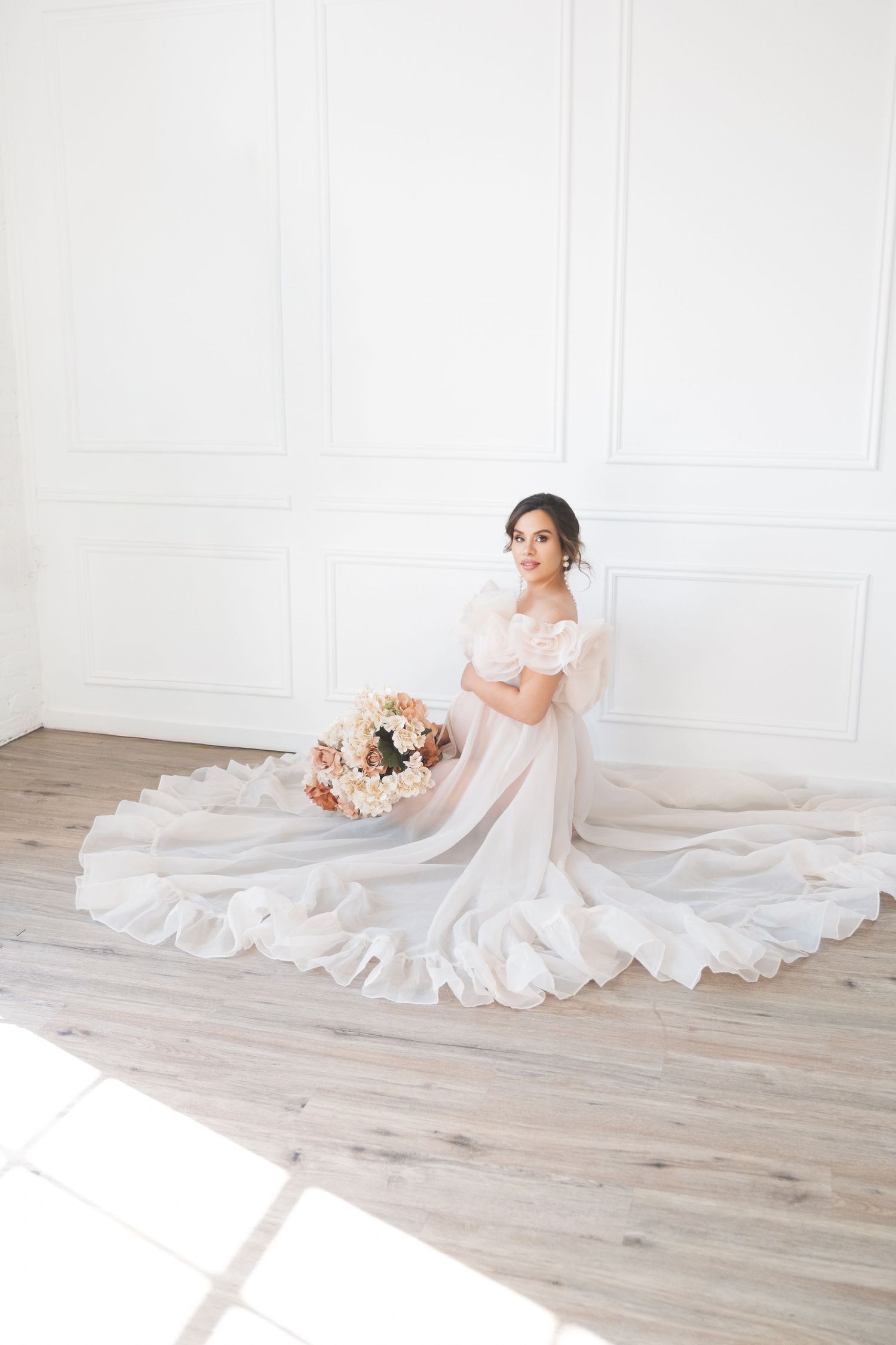 A maternity mum in a flowing white gown poses gracefully on a light floor with pink and peach floral arrangements natural studio, LONDON uk