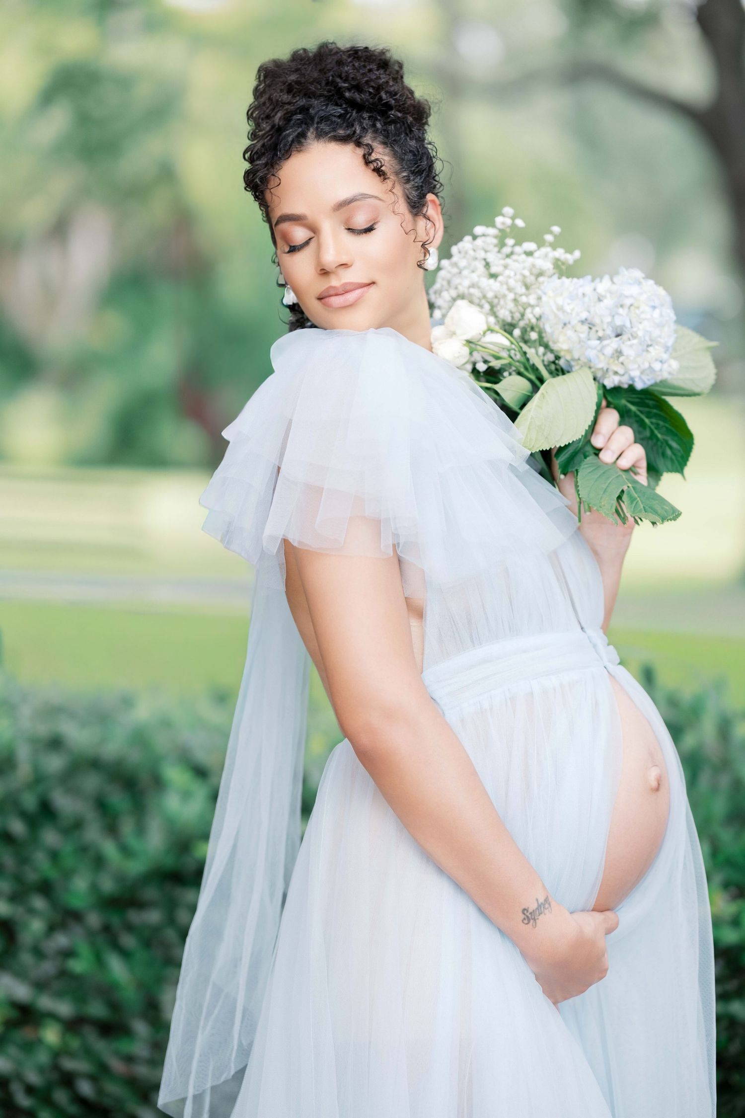 Expectant mother in flowing white dress holds delicate bouquet of white flowers in outdoor garden setting. Motherhood luxury photographer UK