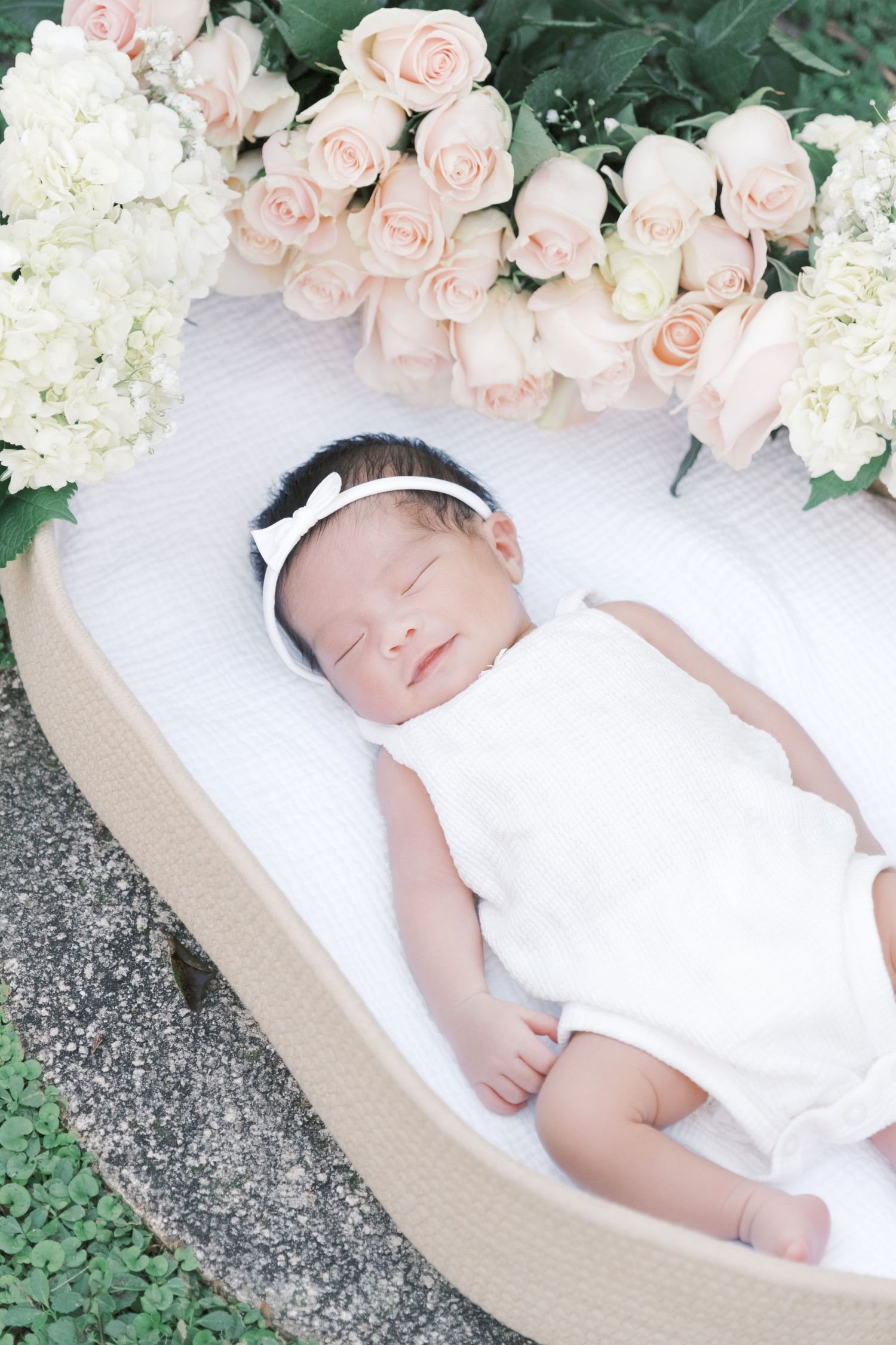 A sleeping newborn baby in white clothing lies peacefully in a woven basket surrounded by delicate pink and white roses. Newborn london uk