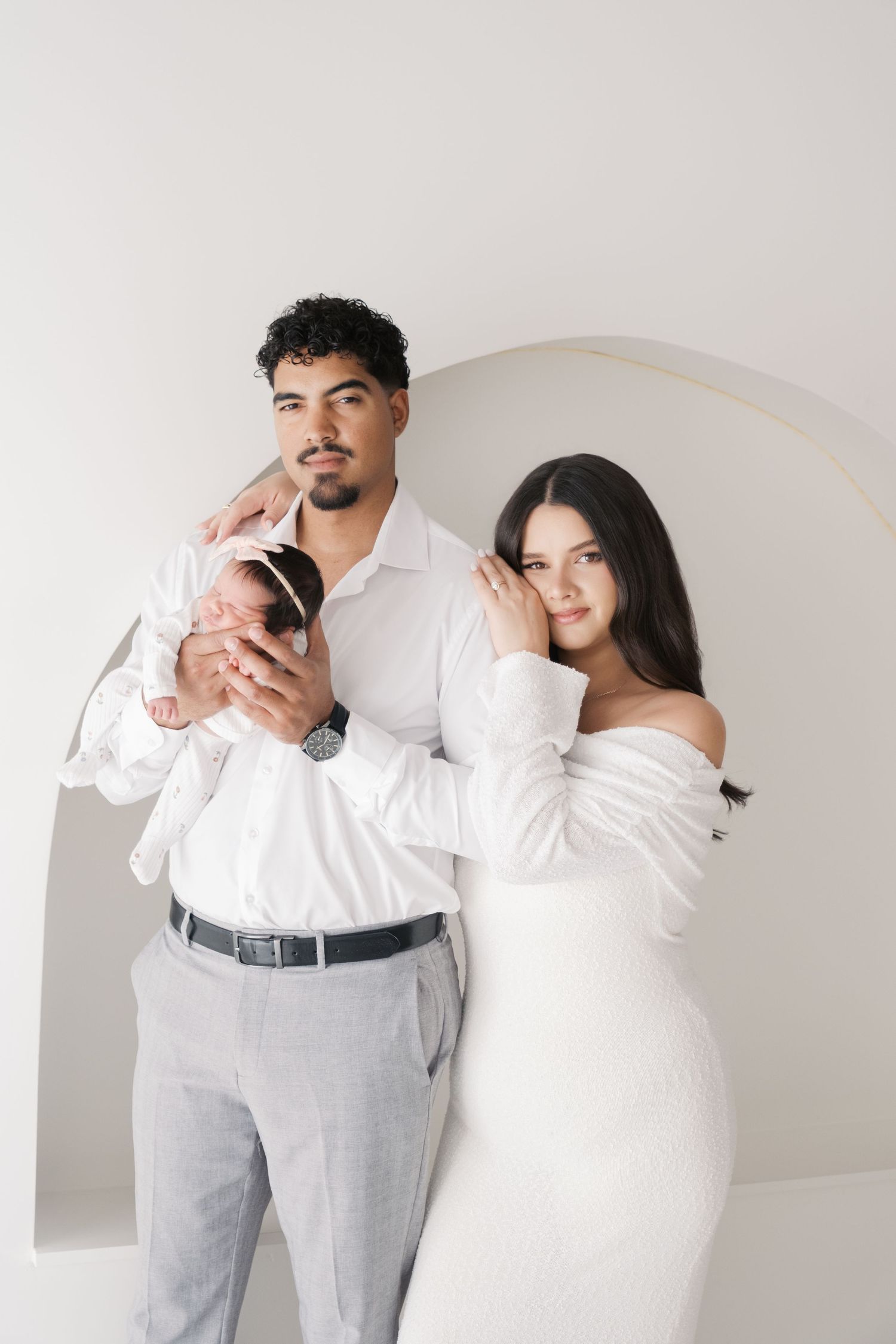 A couple in white outfits poses together against a light gray wall with an arched doorway holding newborn baby. Newborn london and cambridge