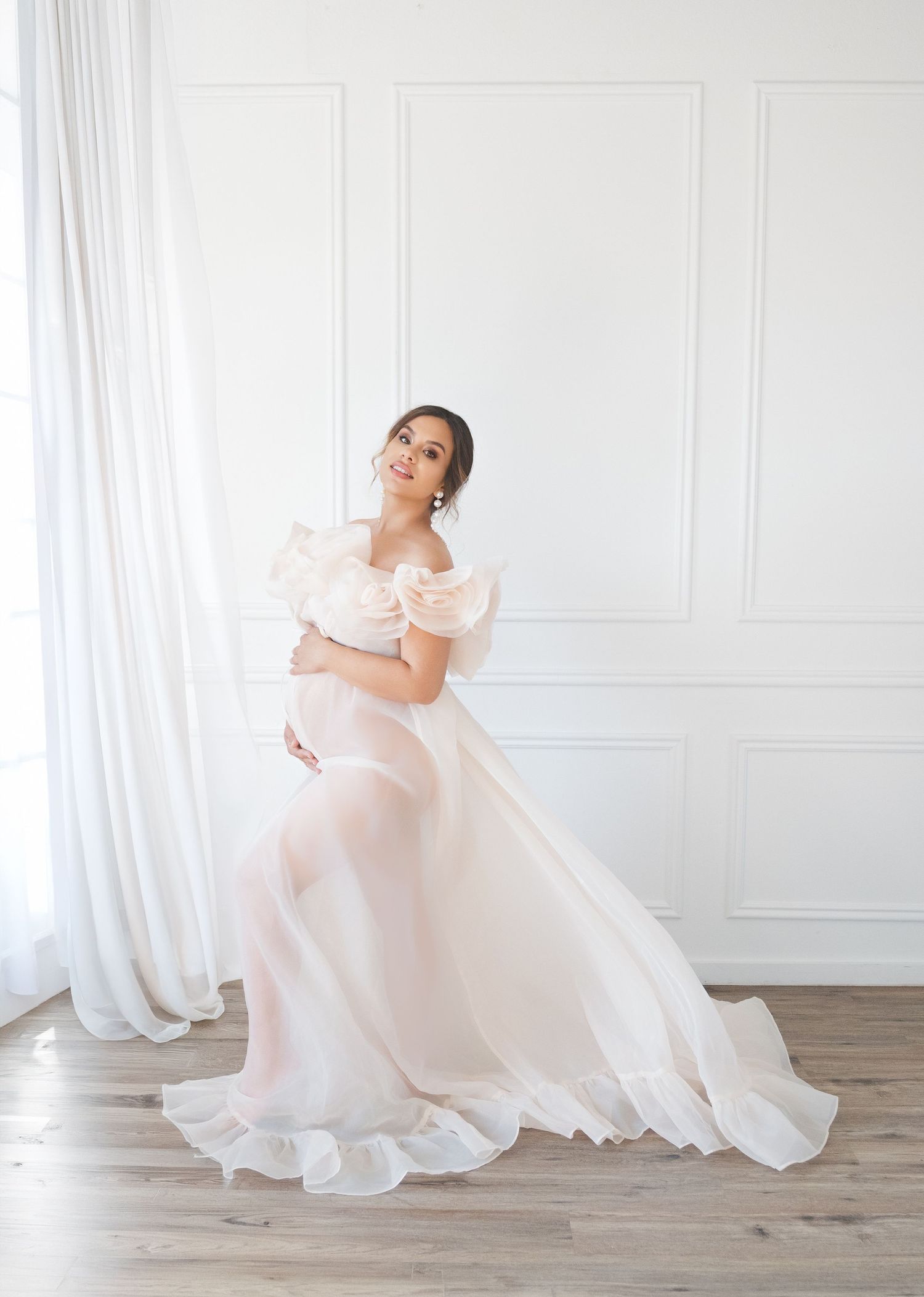 A pregnant mum in a flowing white gown sits gracefully on a light wooden floor holding a pink and peach floral bouquet. London uk sessions