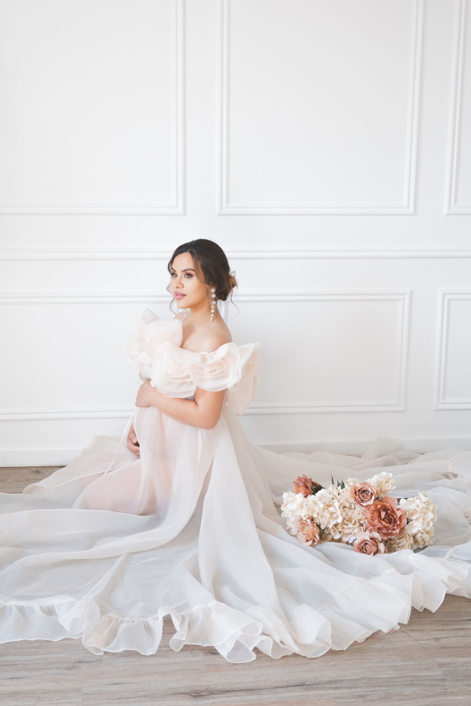 A maternity mum in a flowing white gown poses gracefully on a light floor with pink and peach floral arrangements natural studio, LONDON uk