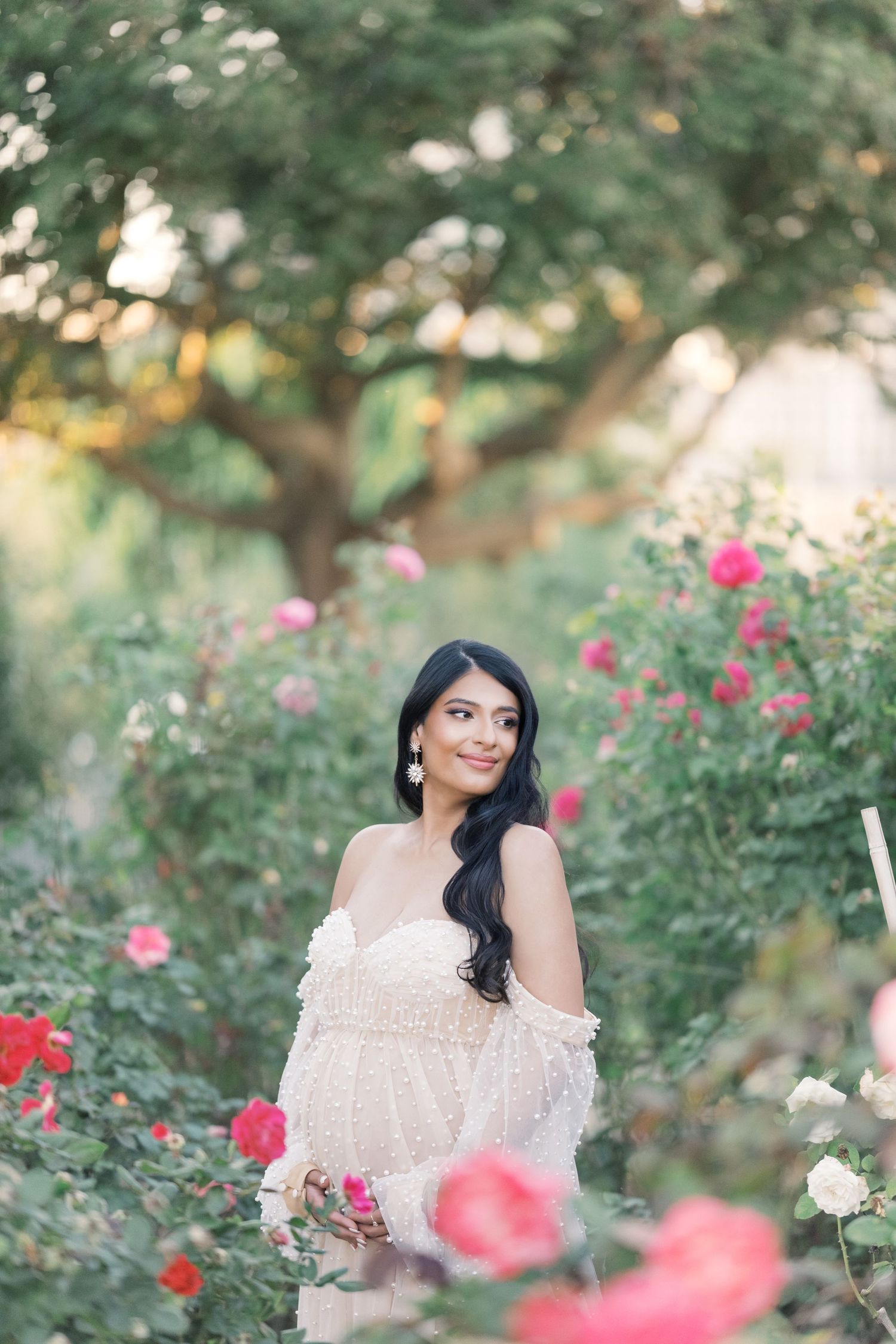 A dreamy portrait in a rose garden with soft bokeh lighting and an off-shoulder white lace dress. Maternity london uk photographer.