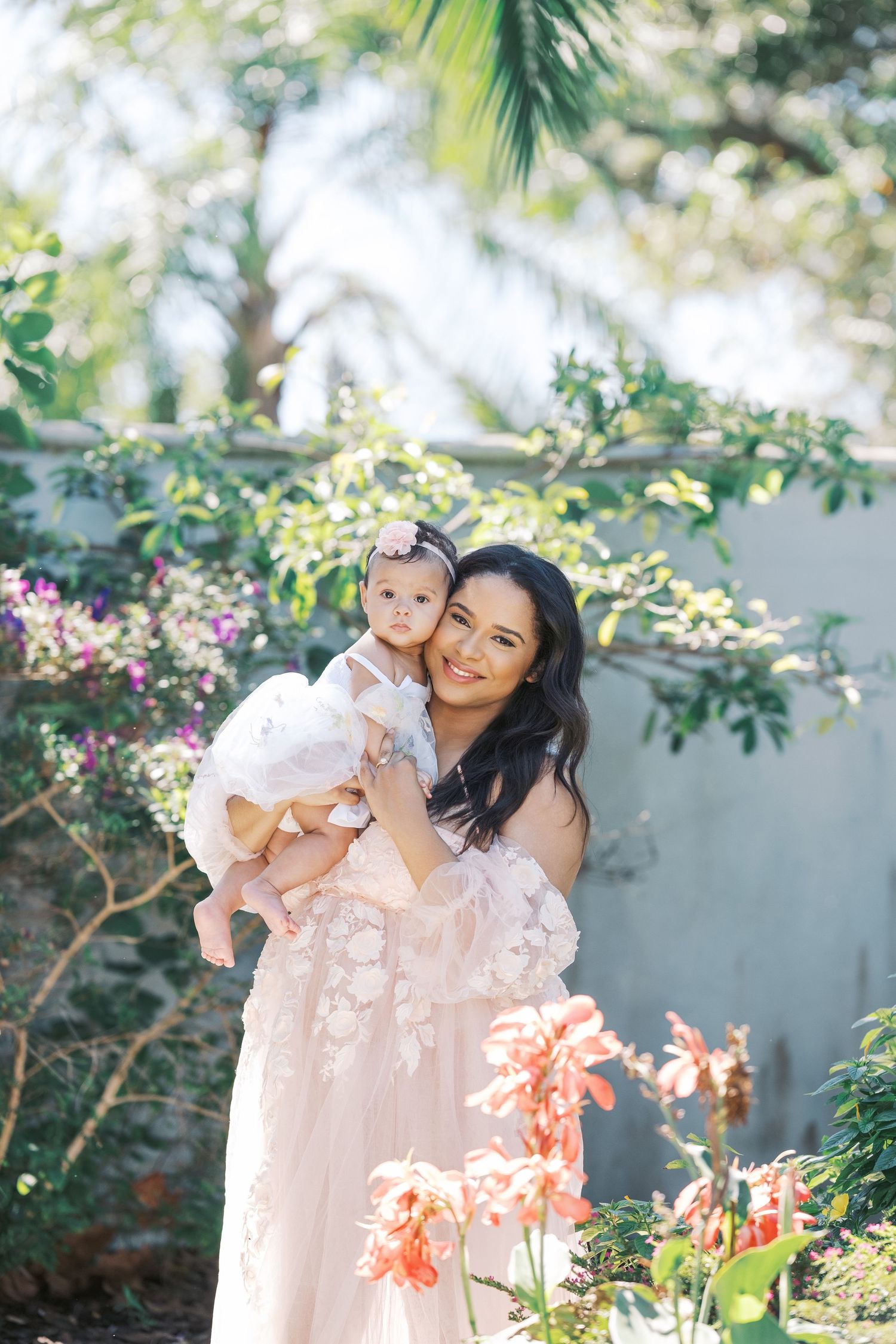 Mother and baby wearing matching pink dresses pose together in a sunny garden with blooming flowers. Motherhood london uk photographer.