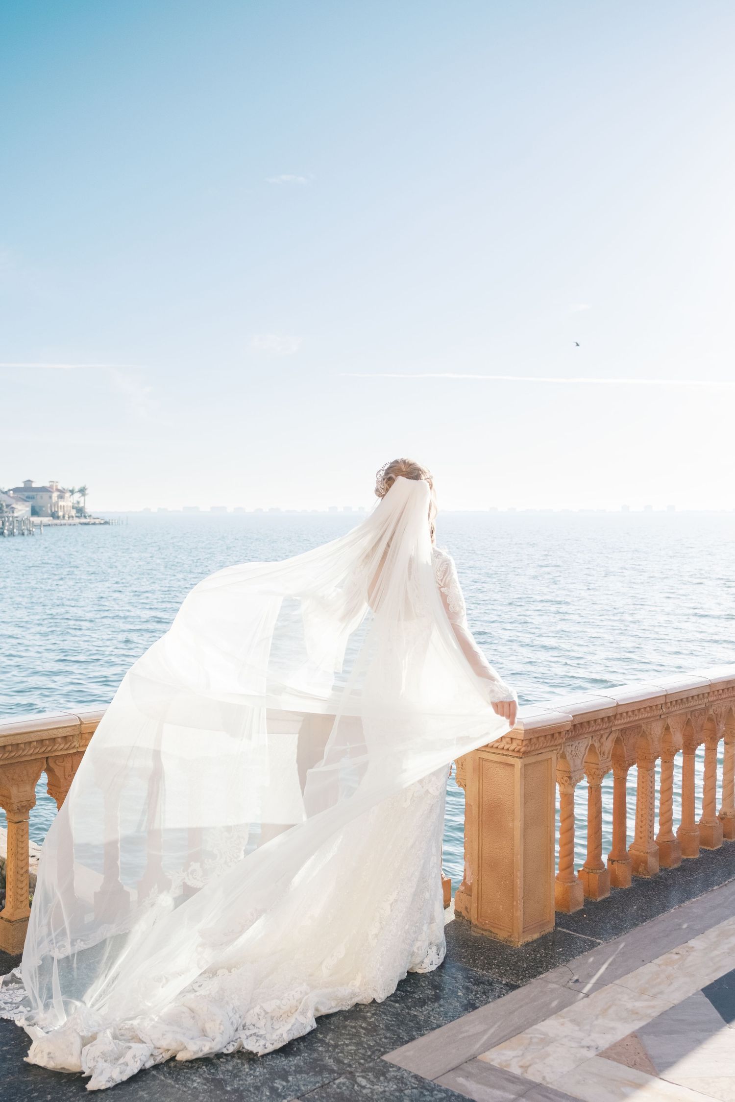 A flowing white wedding dress with a long veil catches the breeze on an oceanfront terrace with wooden railings. cambridge uk photographer