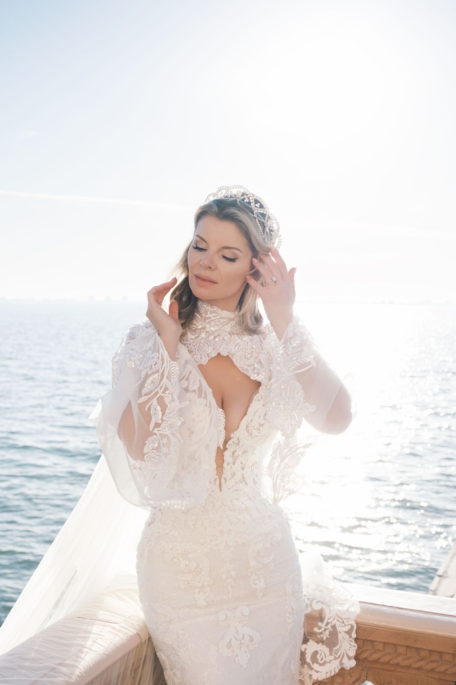 Elegant bridal portrait on a boat with sunlit ocean backdrop featuring a detailed lace wedding gown and tiara. London wedding photographer.