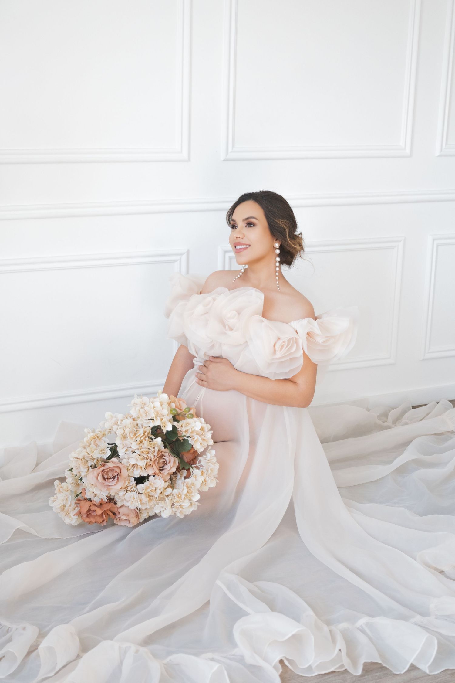 A maternity mum in a flowing white gown poses gracefully on a light floor with pink and peach floral arrangements natural studio, LONDON uk