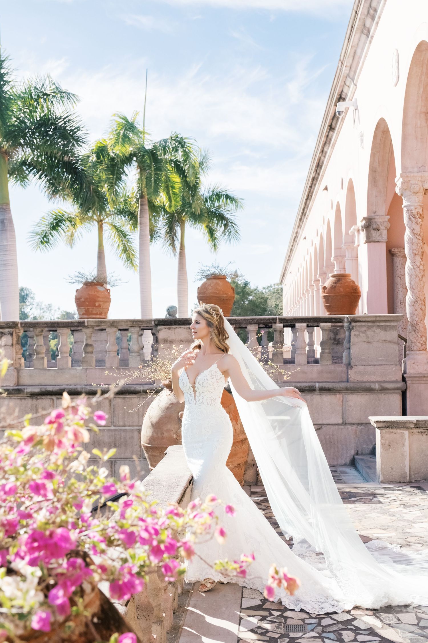 Bride in white wedding dress poses gracefully under Mediterranean-style arches with palm trees and pink flowers. UK wedding photographer