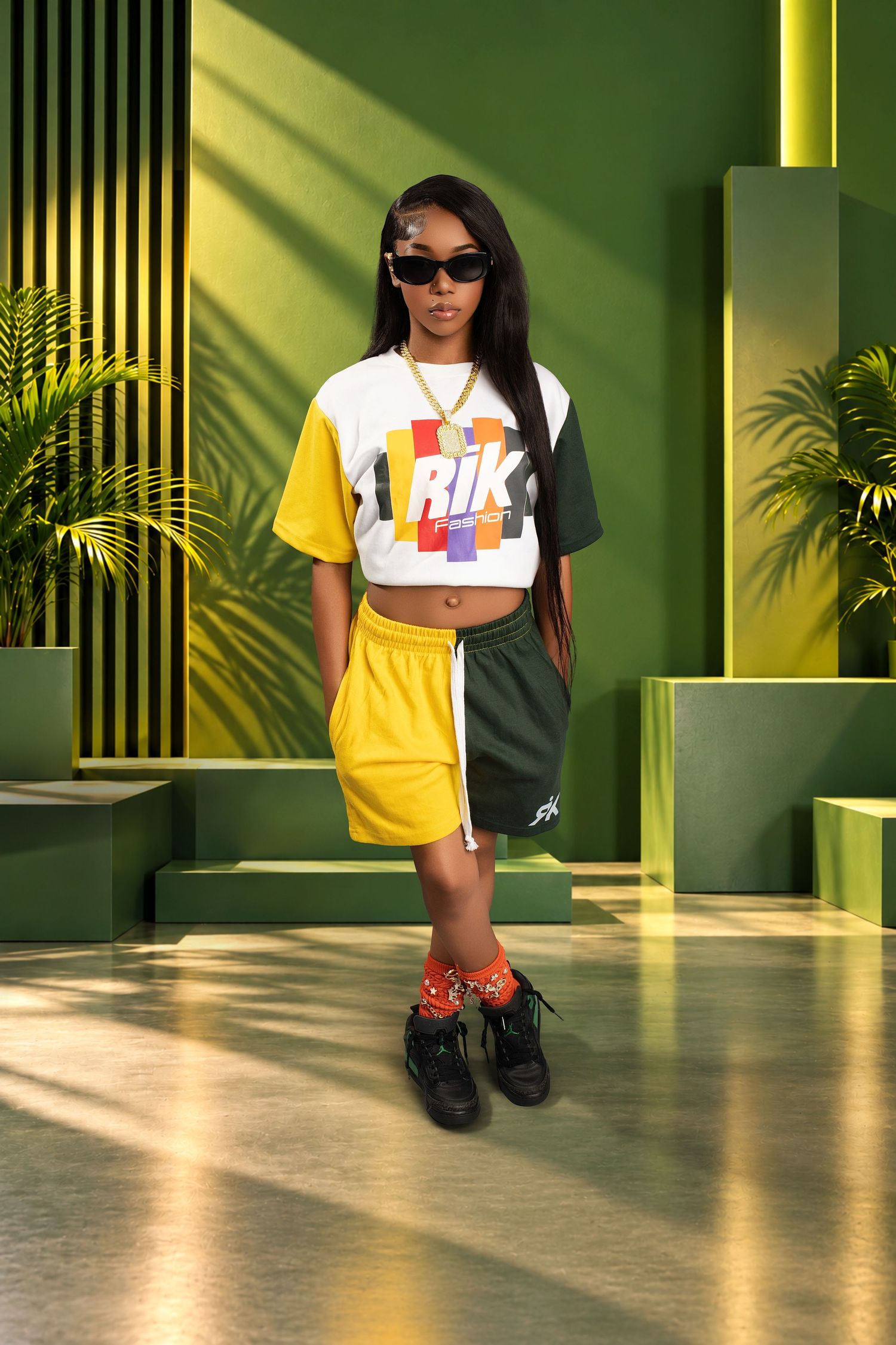 Young girl in sunglasses, a colorful graphic tee, yellow and green shorts and black sneakers posing in a green modern set with palm plants.
