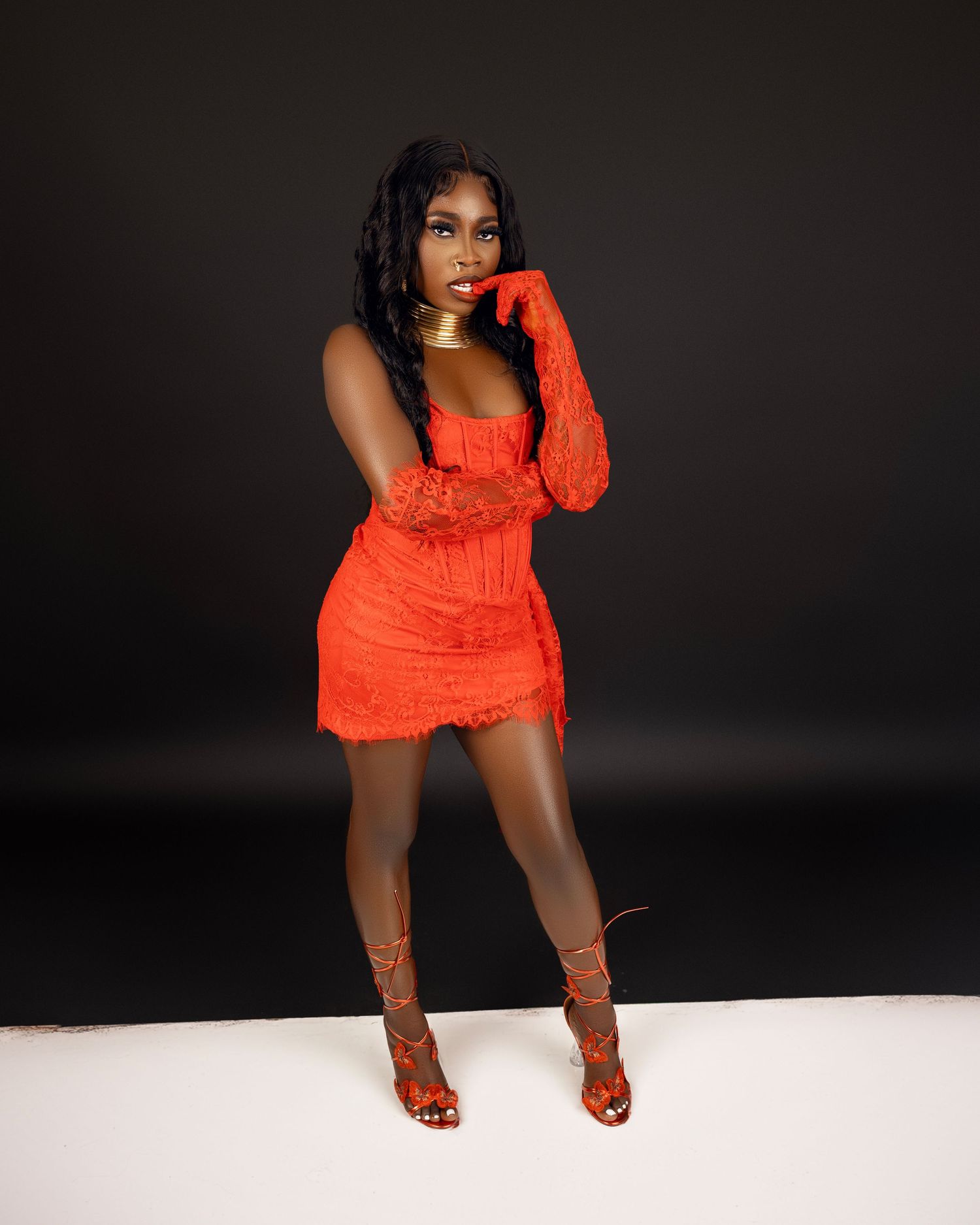 Woman in an orange lace mini dress with matching gloves and lace-up heels poses against a dark studio background.