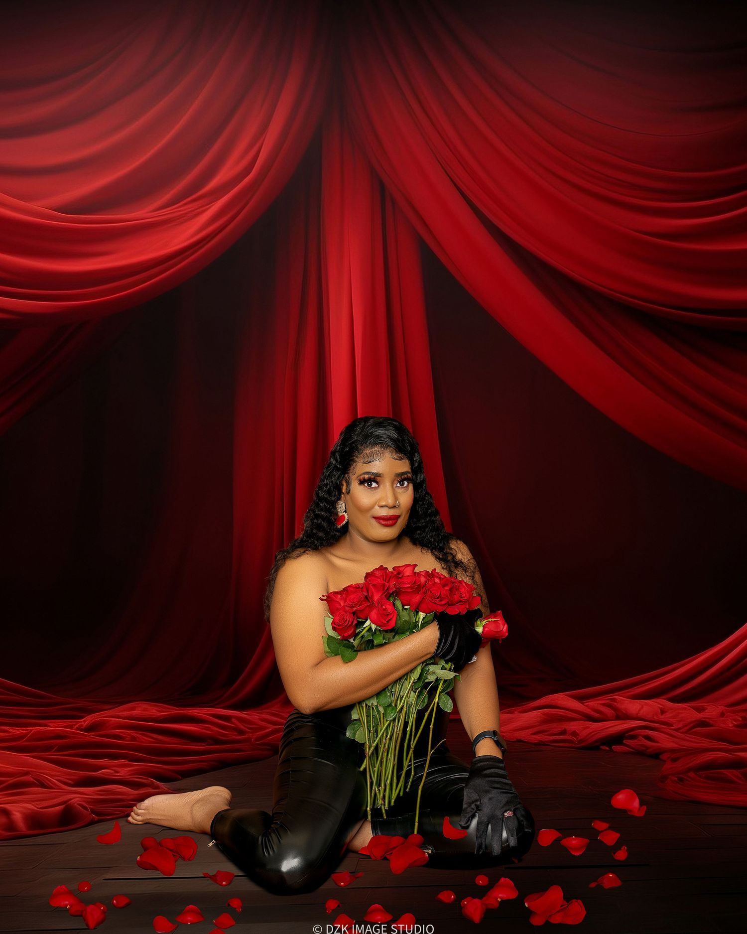Woman in black latex outfit and gloves kneeling on a floor scattered with rose petals, holding red roses before red velvet drapes.