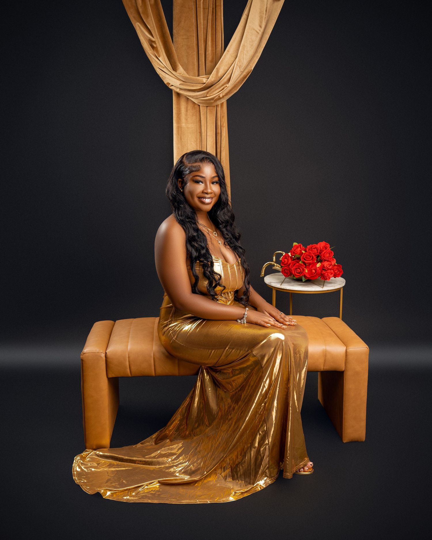 Woman in a gold metallic gown seated on a tan leather bench beside a small table of red roses in a dark studio portrait.