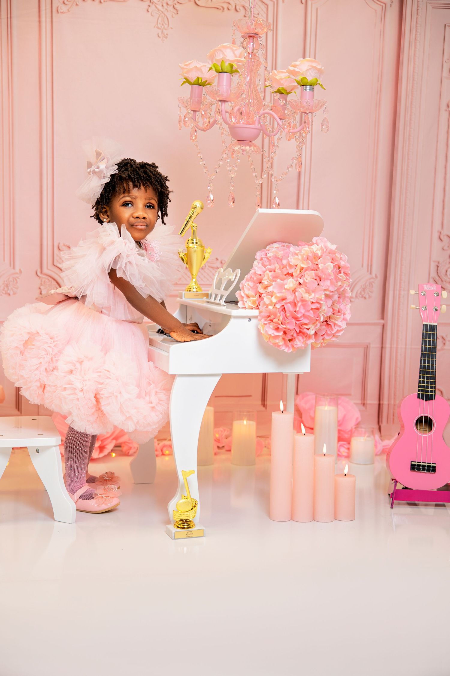 Little girl in a pink tulle dress plays a white toy piano in a pink room with flowers, candles, a chandelier and a ukulele.