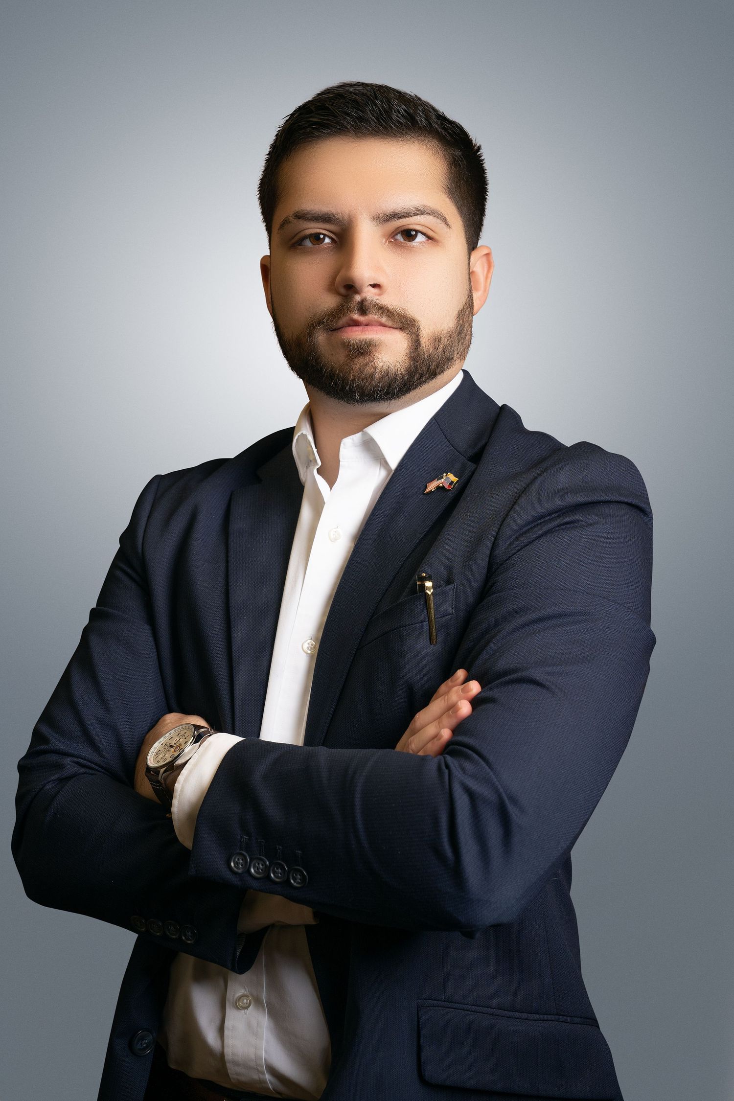 Professional headshot of a bearded man in a navy blazer and white shirt with arms crossed against a gray studio backdrop.