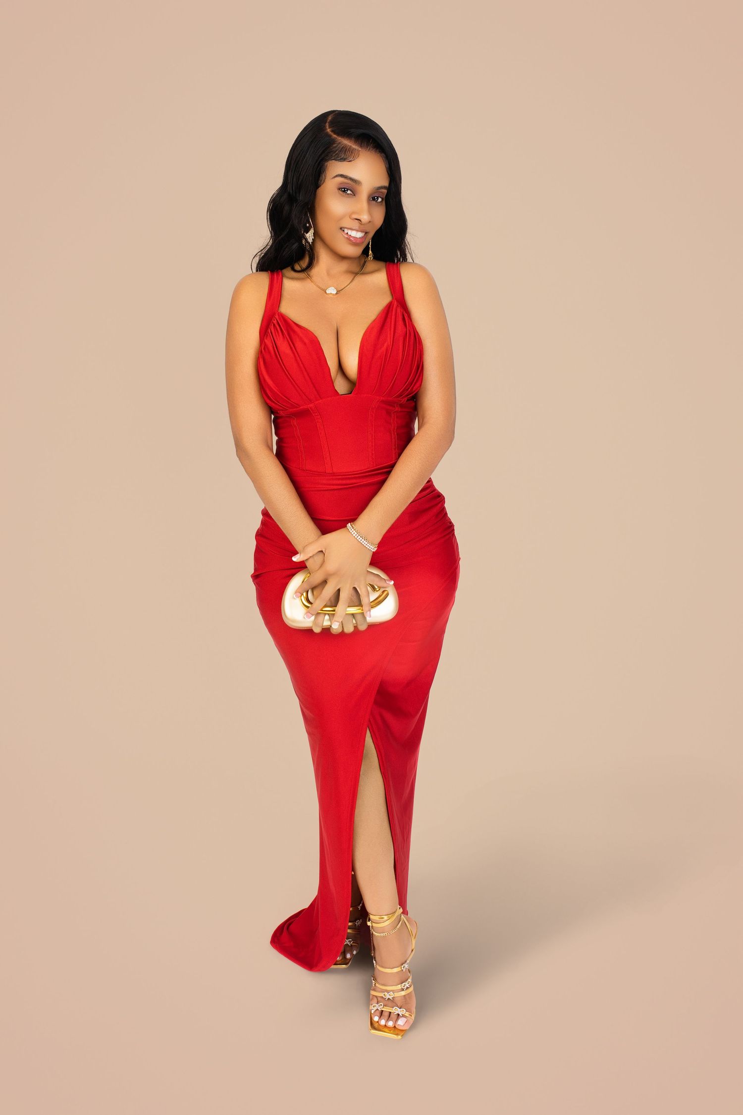 Woman in a red draped gown with a high slit holding a gold clutch against a beige studio backdrop.