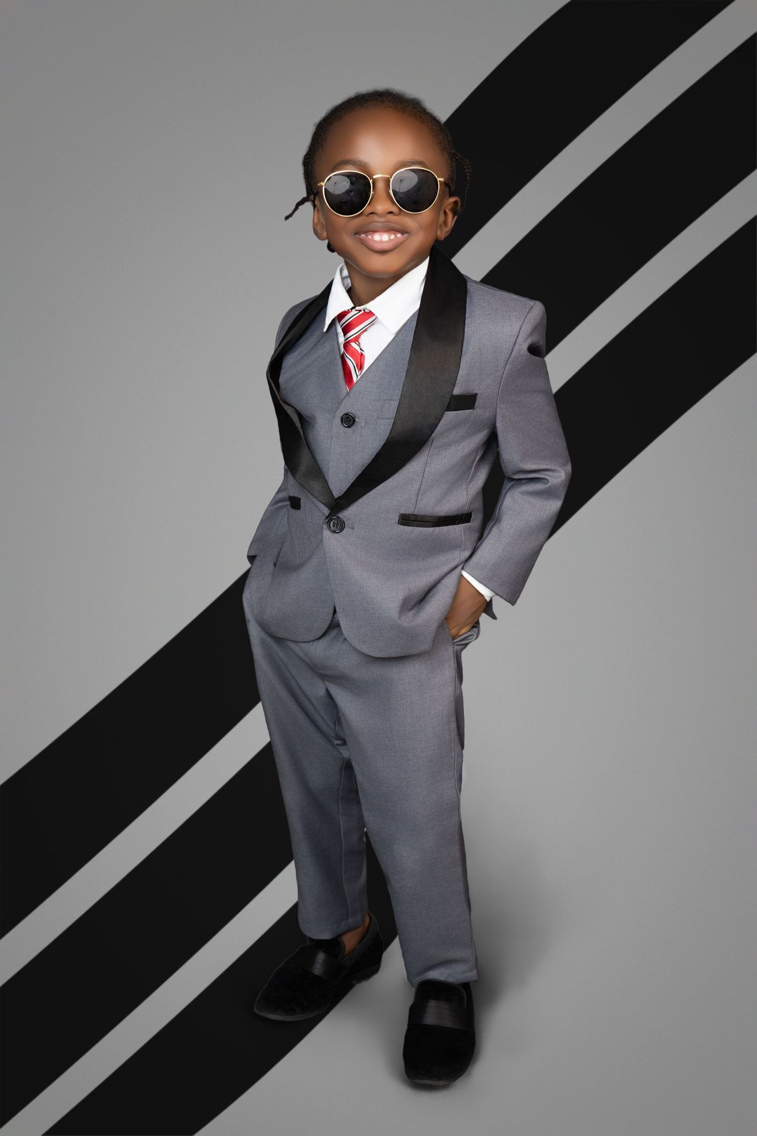 Smiling young boy in round sunglasses, gray tuxedo suit and red striped tie poses with hands in pockets against a striped backdrop.