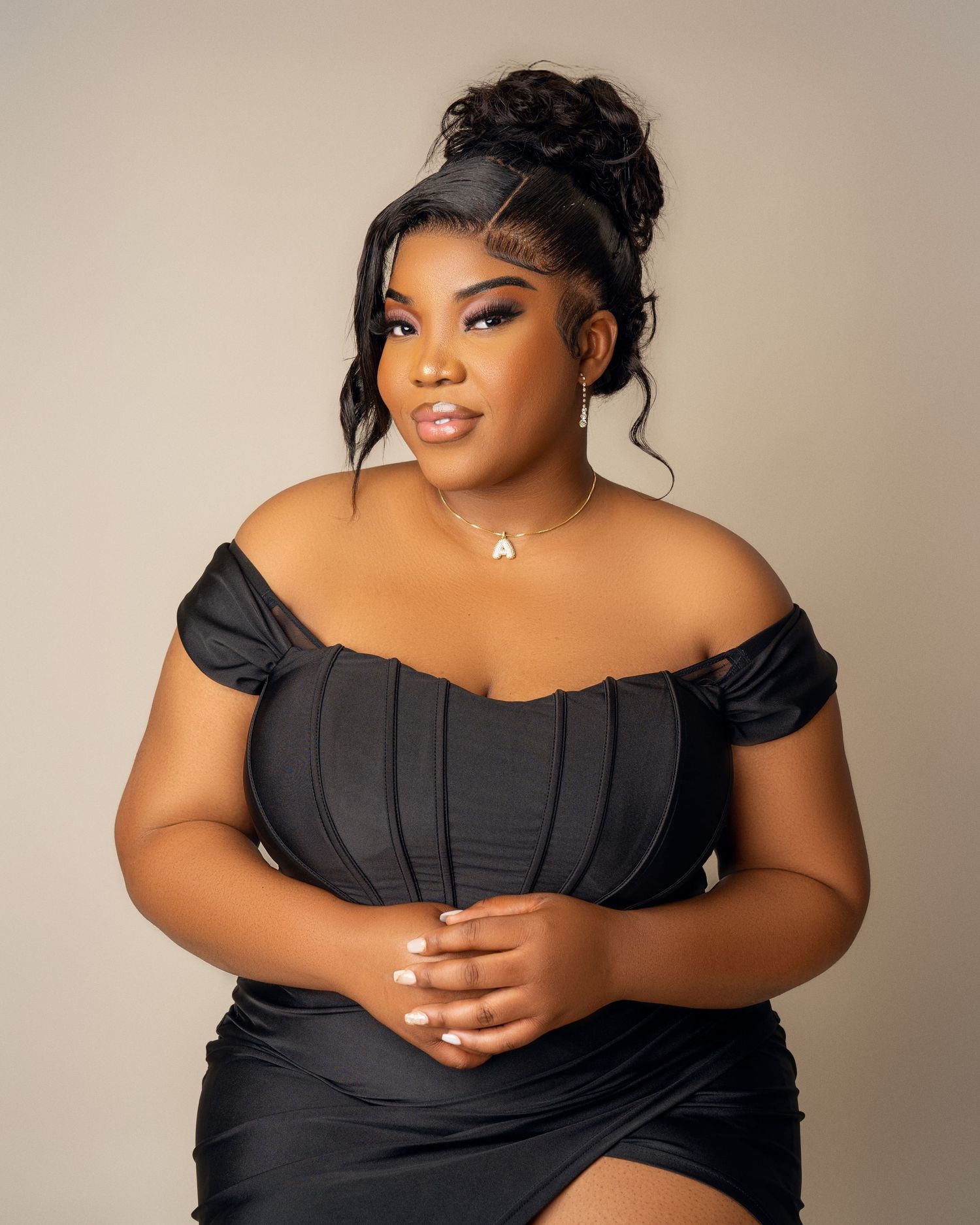 Woman in a black off-shoulder corset dress with an updo and pendant necklace posing against a soft neutral studio background.
