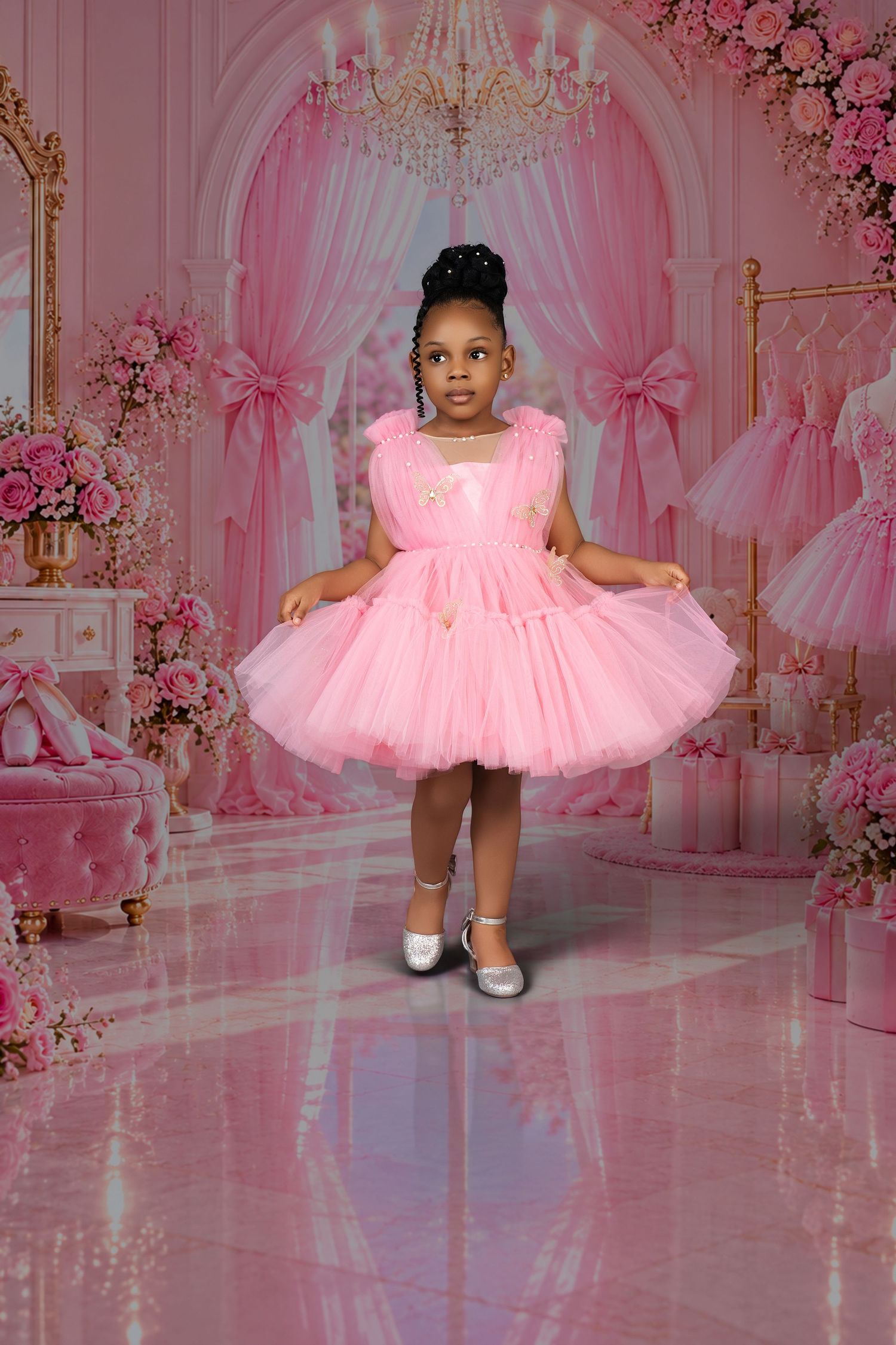 Little girl in a pink tulle party dress and silver shoes stands in a pink room with chandelier, floral arrangements and bow curtains.