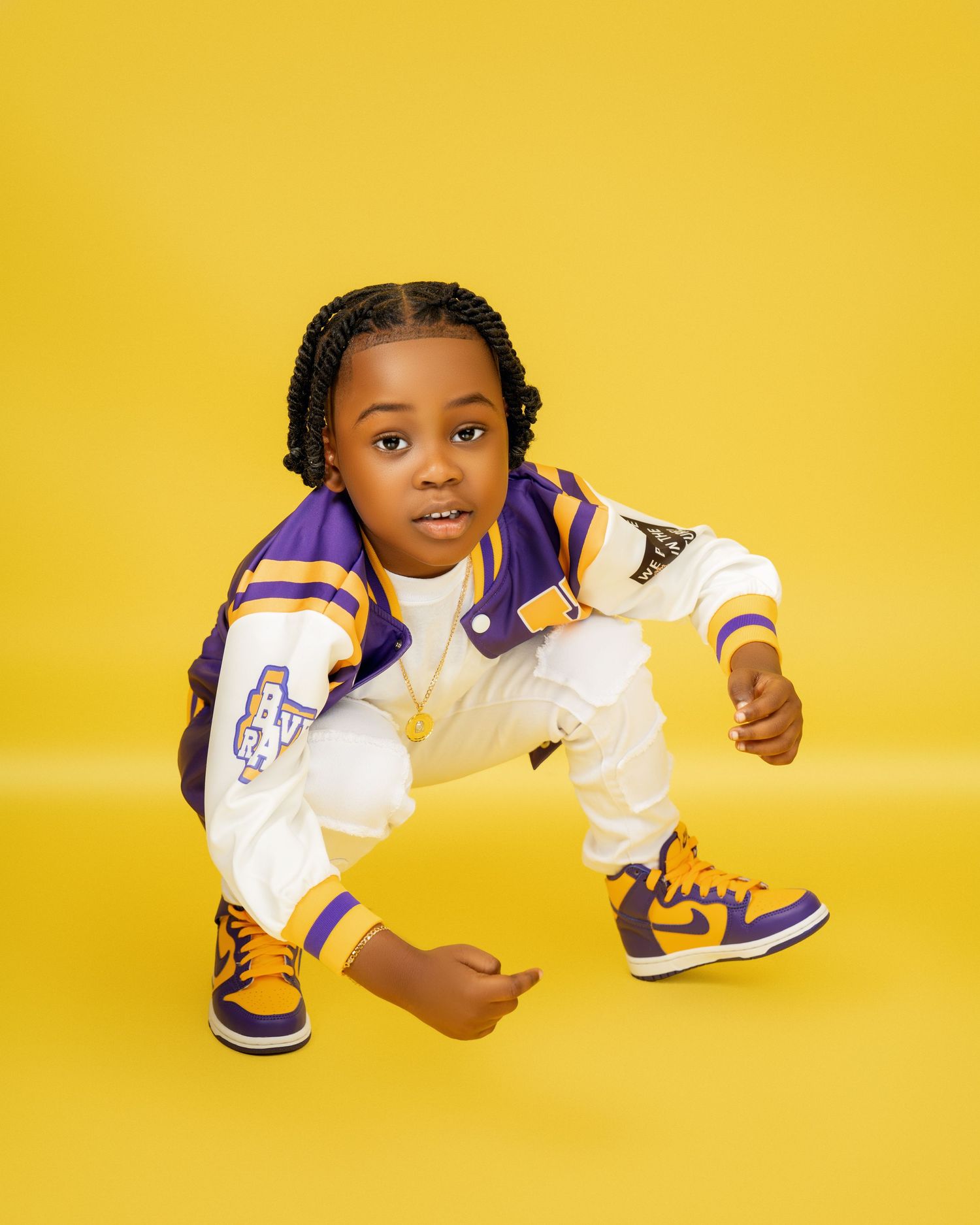 Boy with braids crouches in a purple and white varsity jacket and matching sneakers against a bright yellow background.