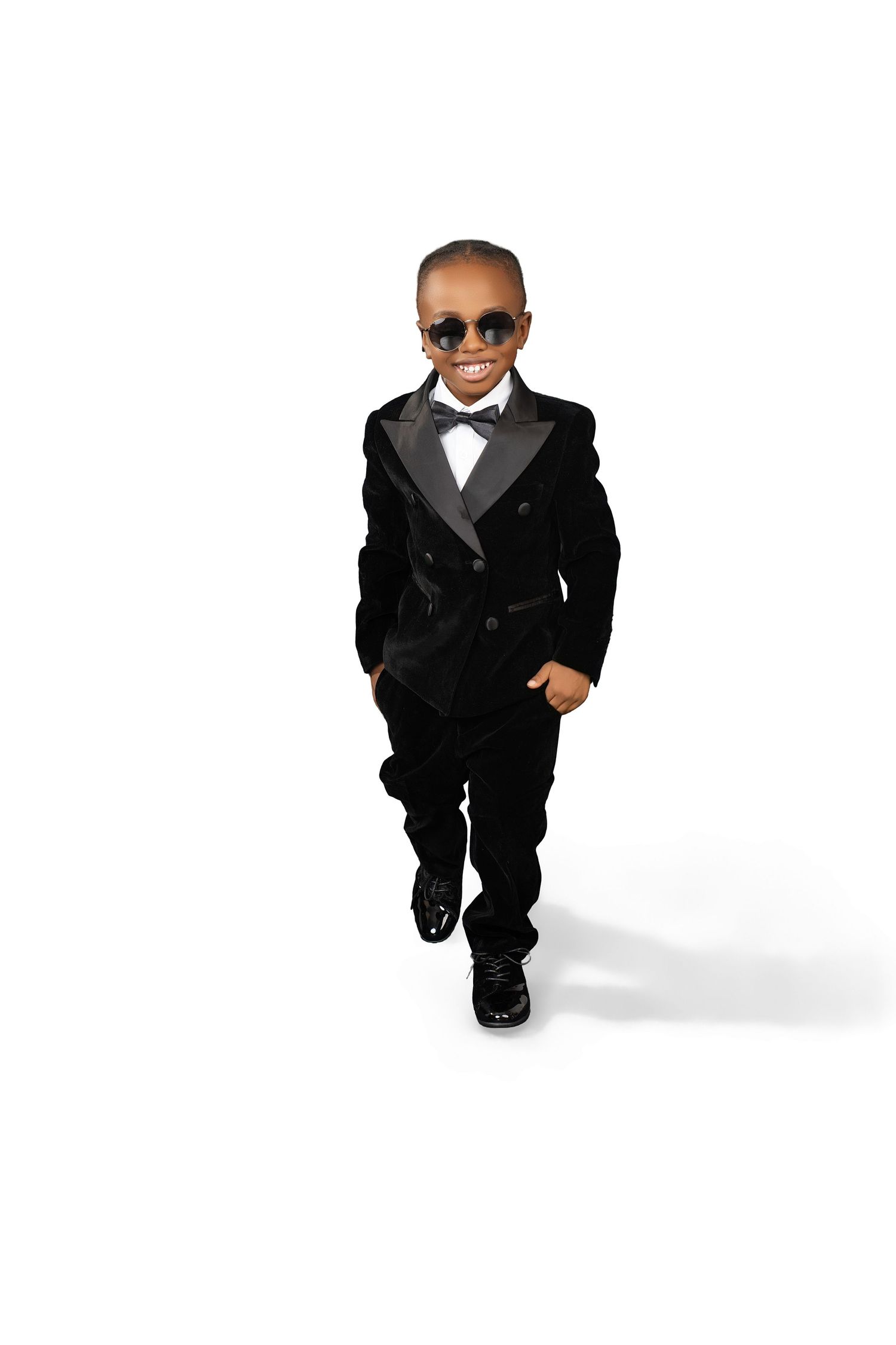 Boy in sunglasses, a black velvet tuxedo jacket, bow tie and polished shoes walks confidently against a white background.