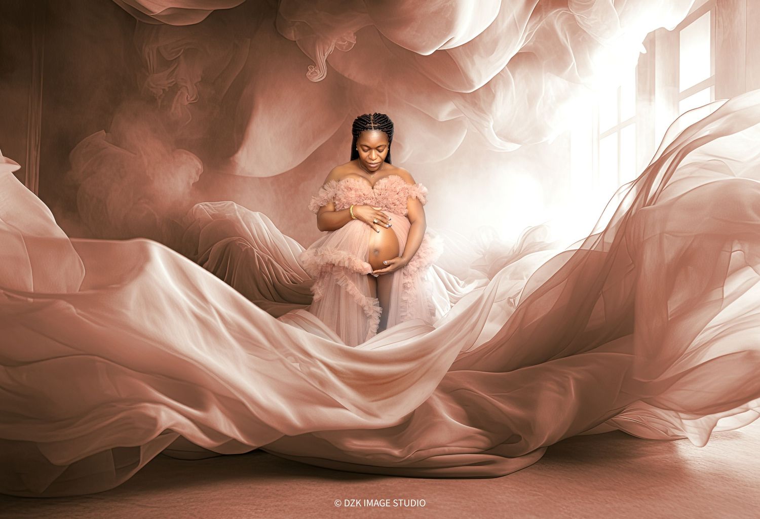 Pregnant woman in a pink ruffled top sits in a swirl of billowing blush fabric in an artistic maternity portrait with soft light.