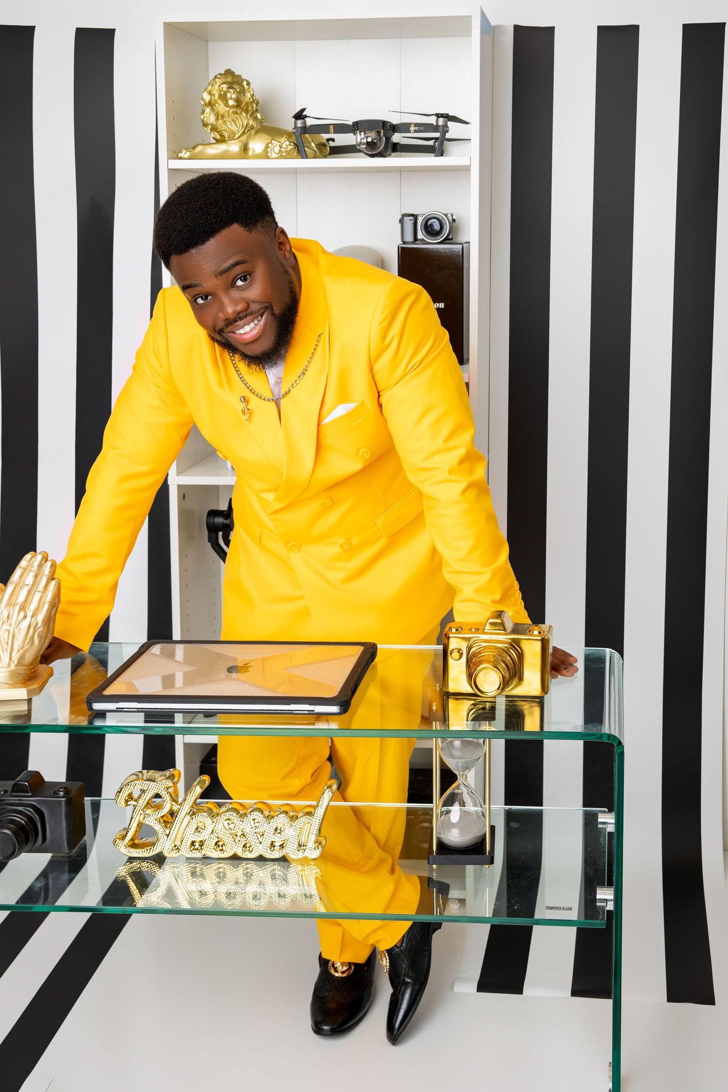 Smiling man in a bright yellow suit leaning over a glass table with gold cameras, a Blessed sign and a striped backdrop.