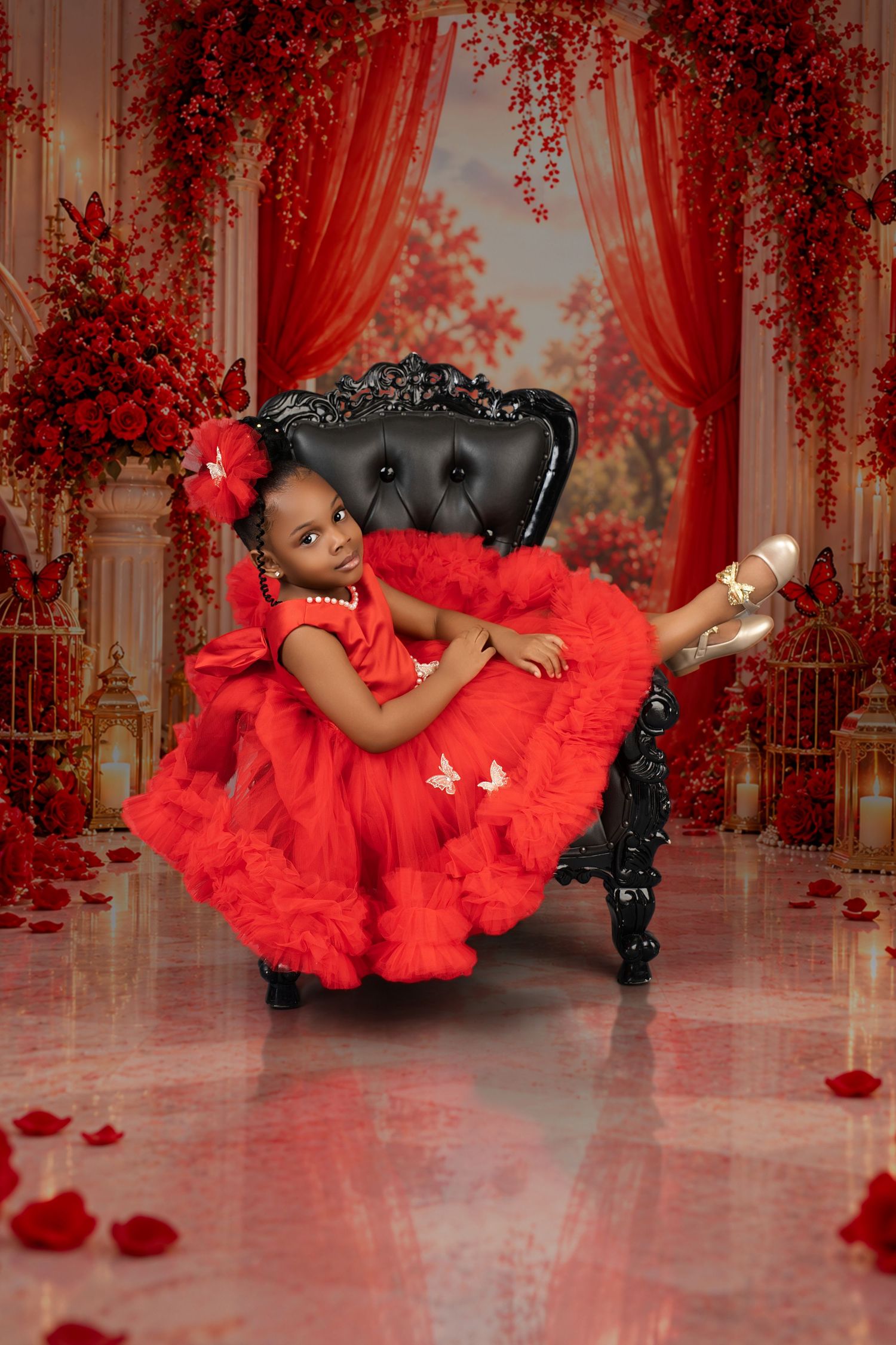 Girl in a ruffled red tulle dress and red hair bow reclines on a black tufted chair surrounded by red roses, butterflies and rose petals.