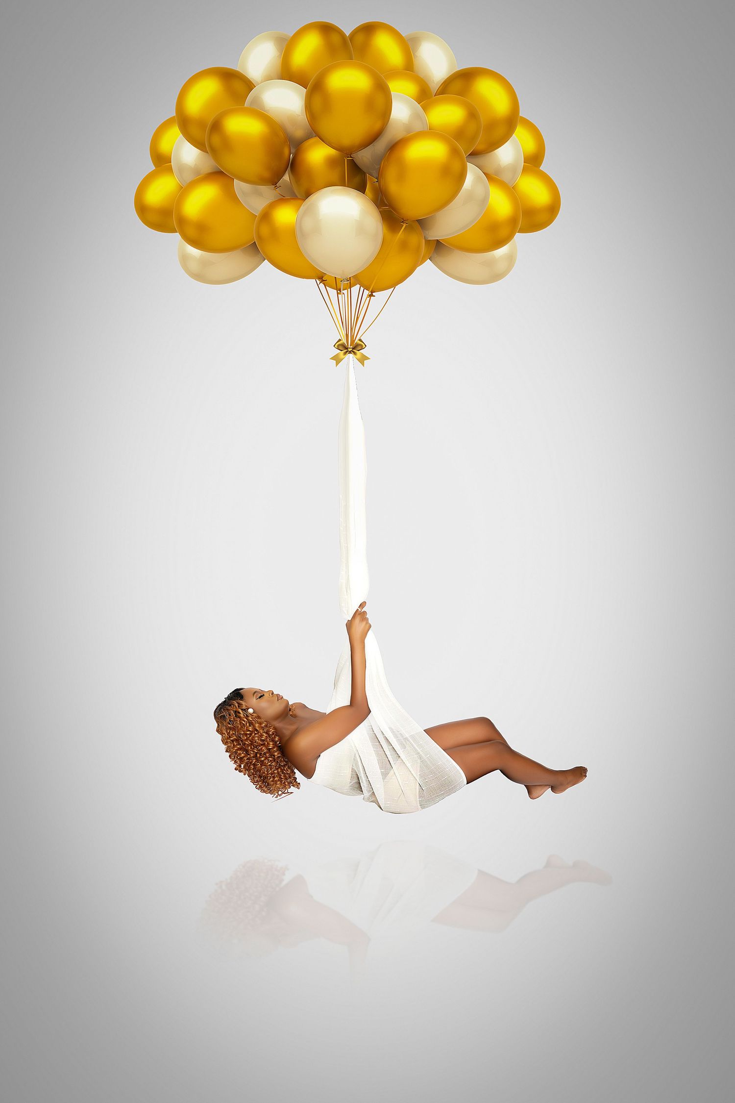 Woman in a white dress holds a ribbon tied to a bunch of gold and cream balloons as she floats above a soft reflective floor.