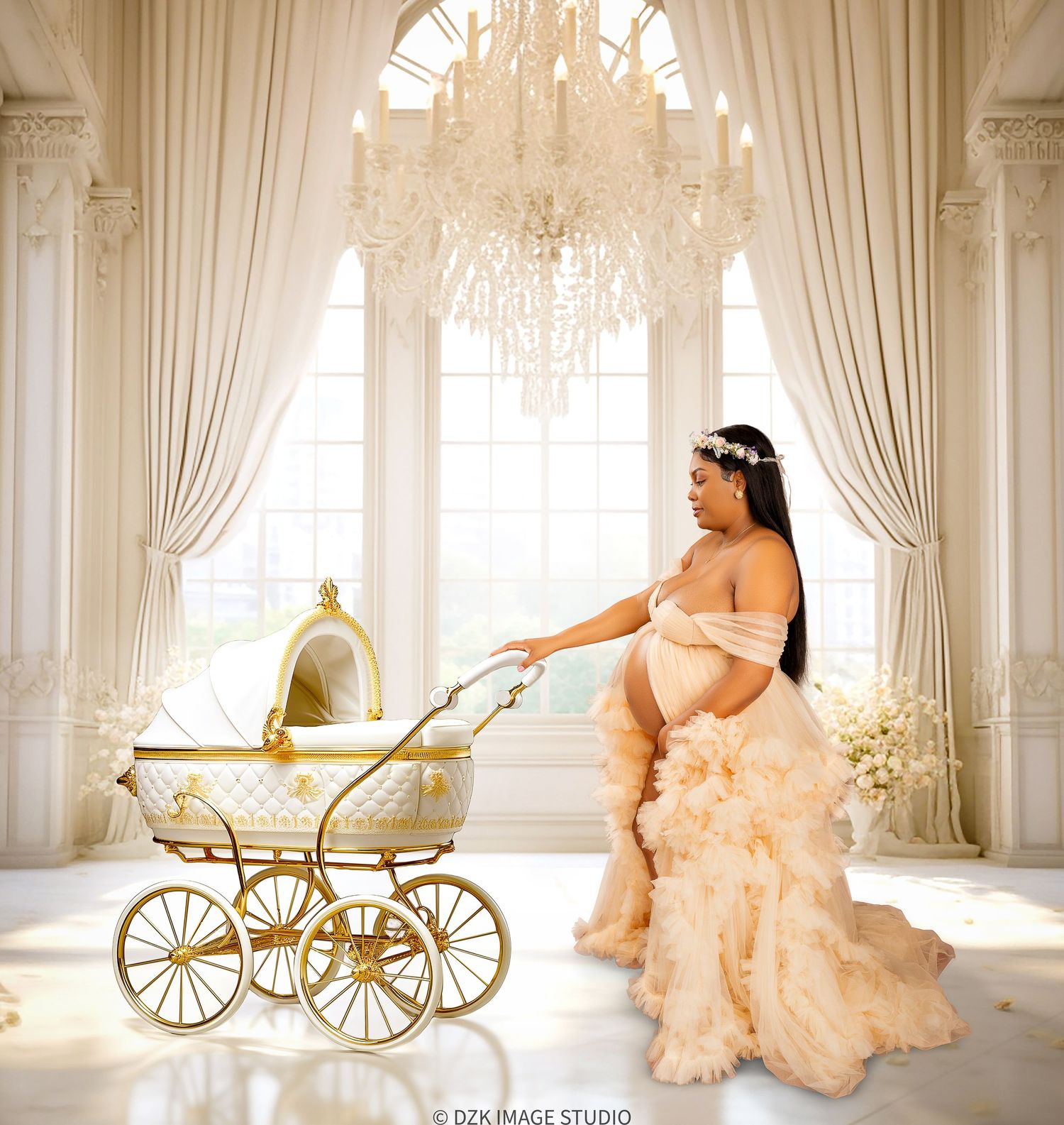Pregnant woman in a cream tulle ruffled gown and floral crown beside a gold vintage baby carriage in an elegant chandelier-lit room.