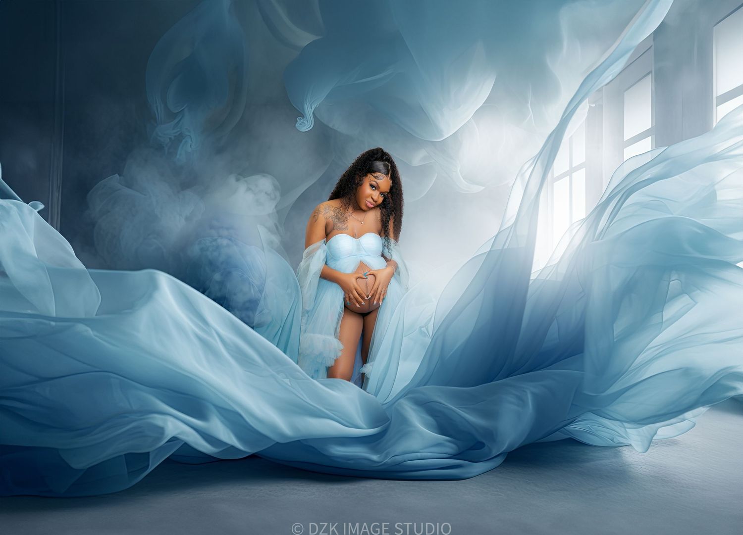 Pregnant woman in a light blue outfit stands amid flowing blue fabric and misty smoke in a dreamy maternity photoshoot.