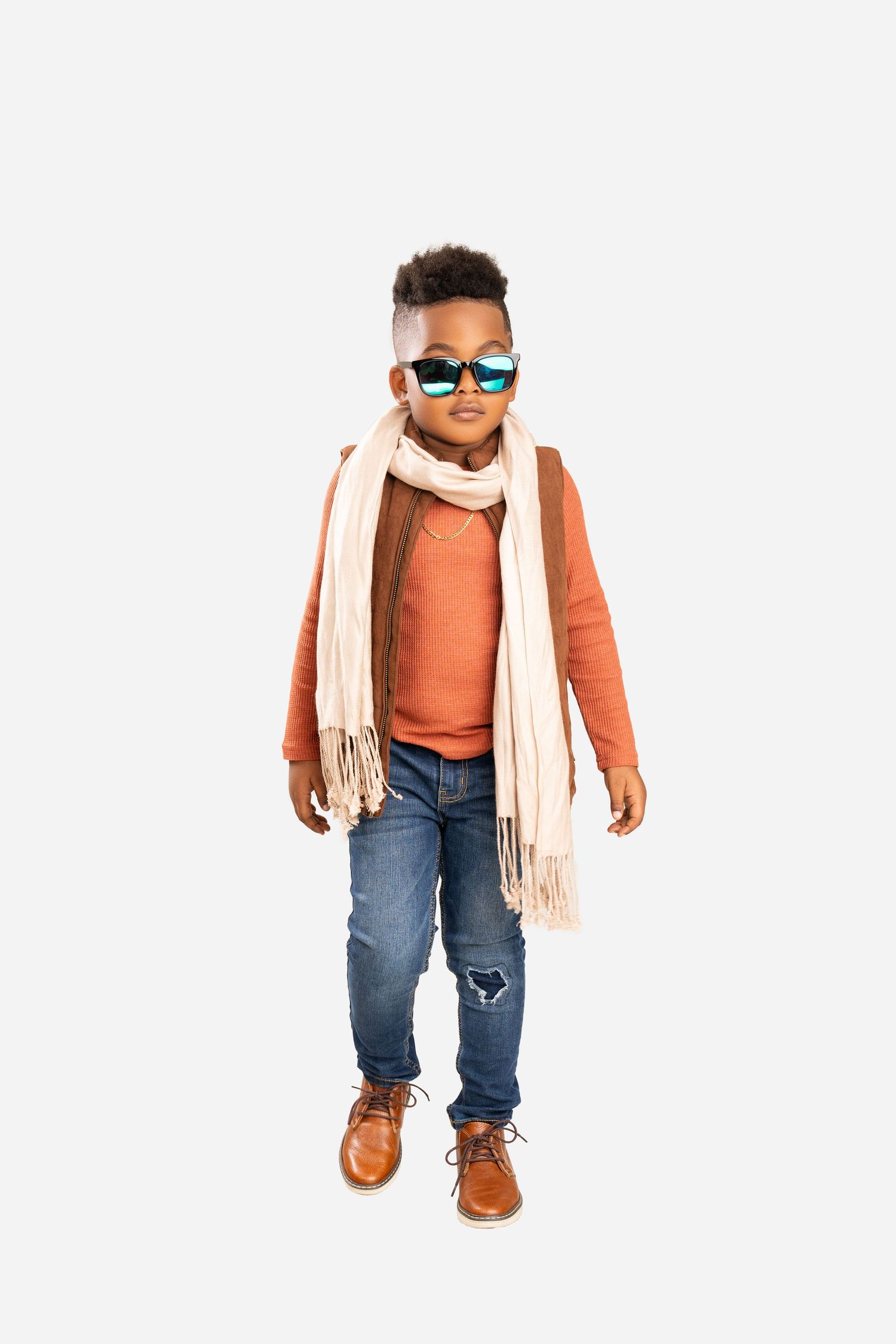 Boy in an orange sweater, cream fringe scarf, ripped jeans, brown boots and teal mirrored sunglasses standing on a white background.