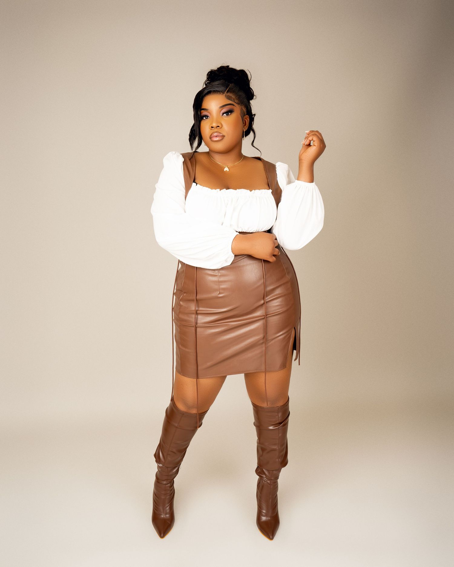 Woman in a white puff-sleeve top, brown leather skirt, and knee-high brown boots posing in a studio with a neutral backdrop.