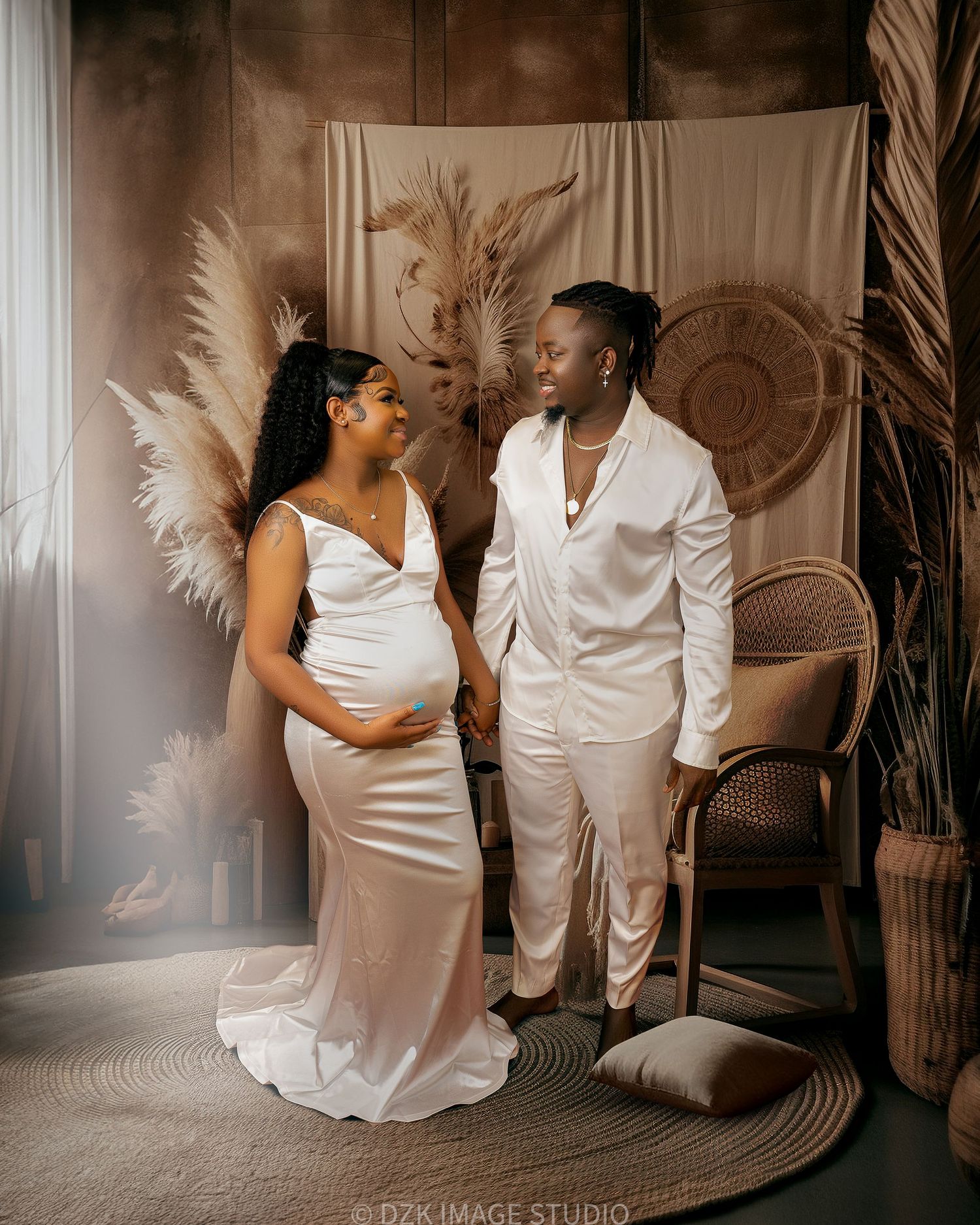 Expectant couple in matching white satin outfits pose in a boho studio with pampas grass, a woven wall piece and a rattan chair.