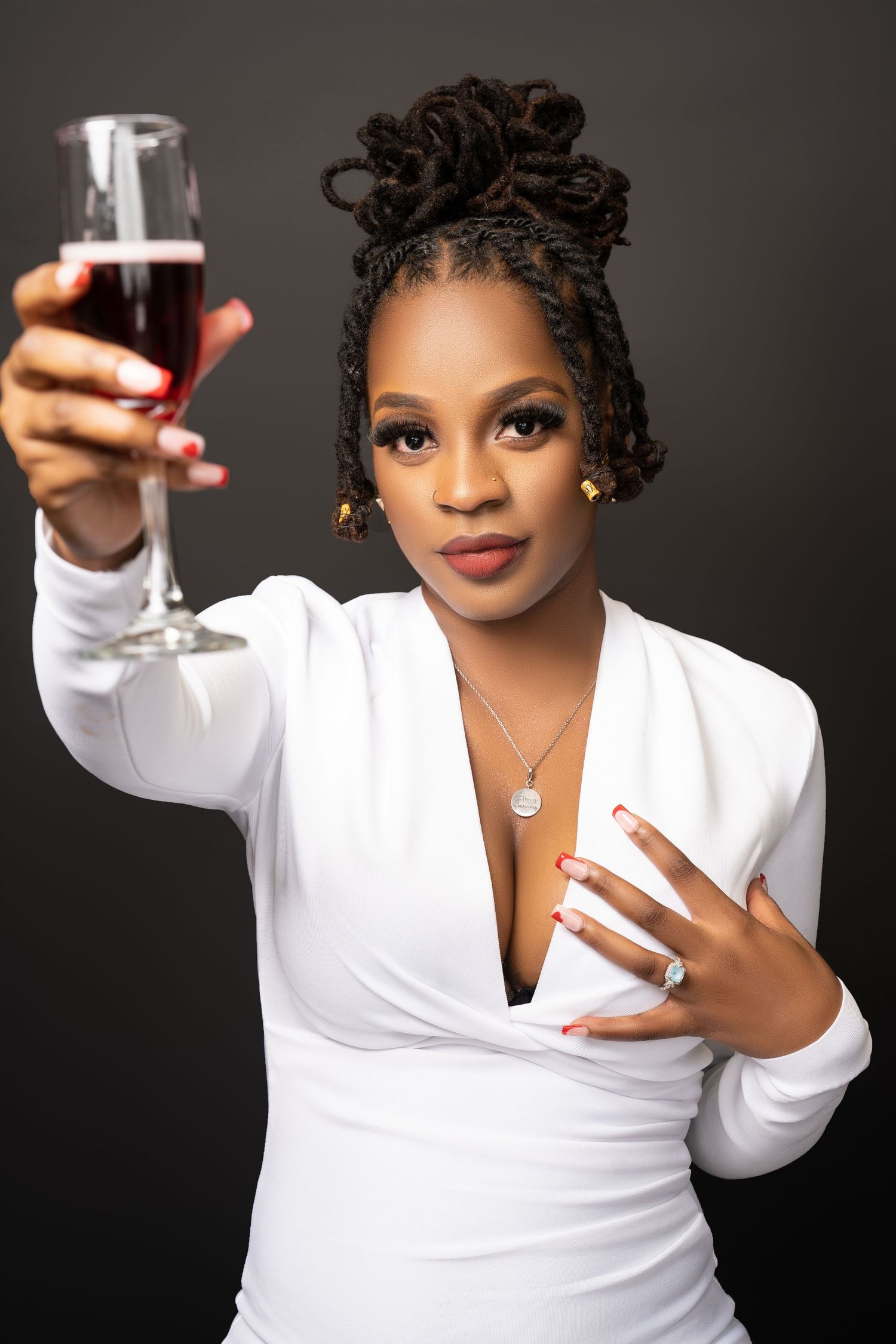 Woman in a white wrap dress raises a glass of red wine in a toast against a dark studio background.