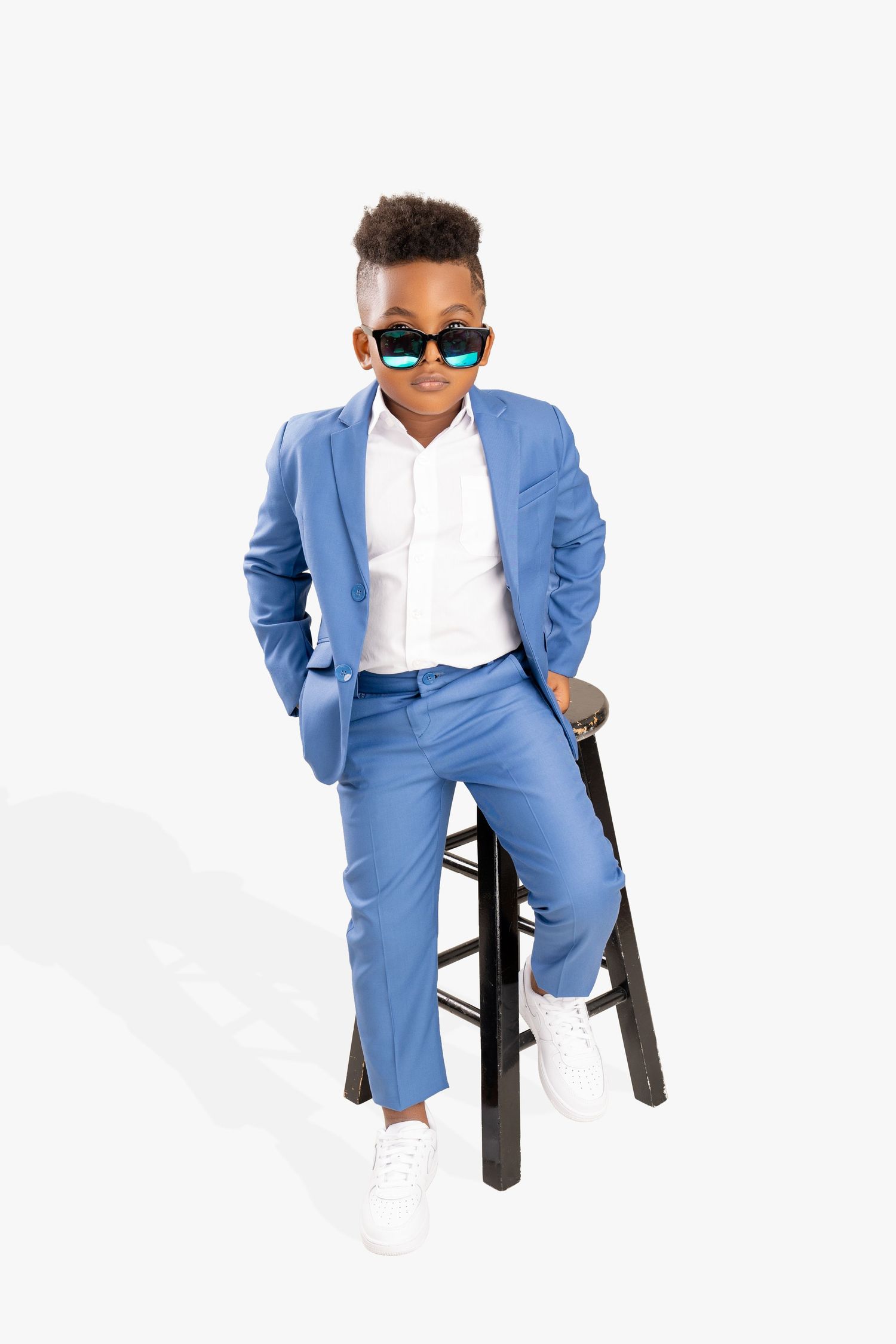 Stylish boy in a blue suit, white shirt, green mirrored sunglasses and white sneakers sitting on a black wooden stool.