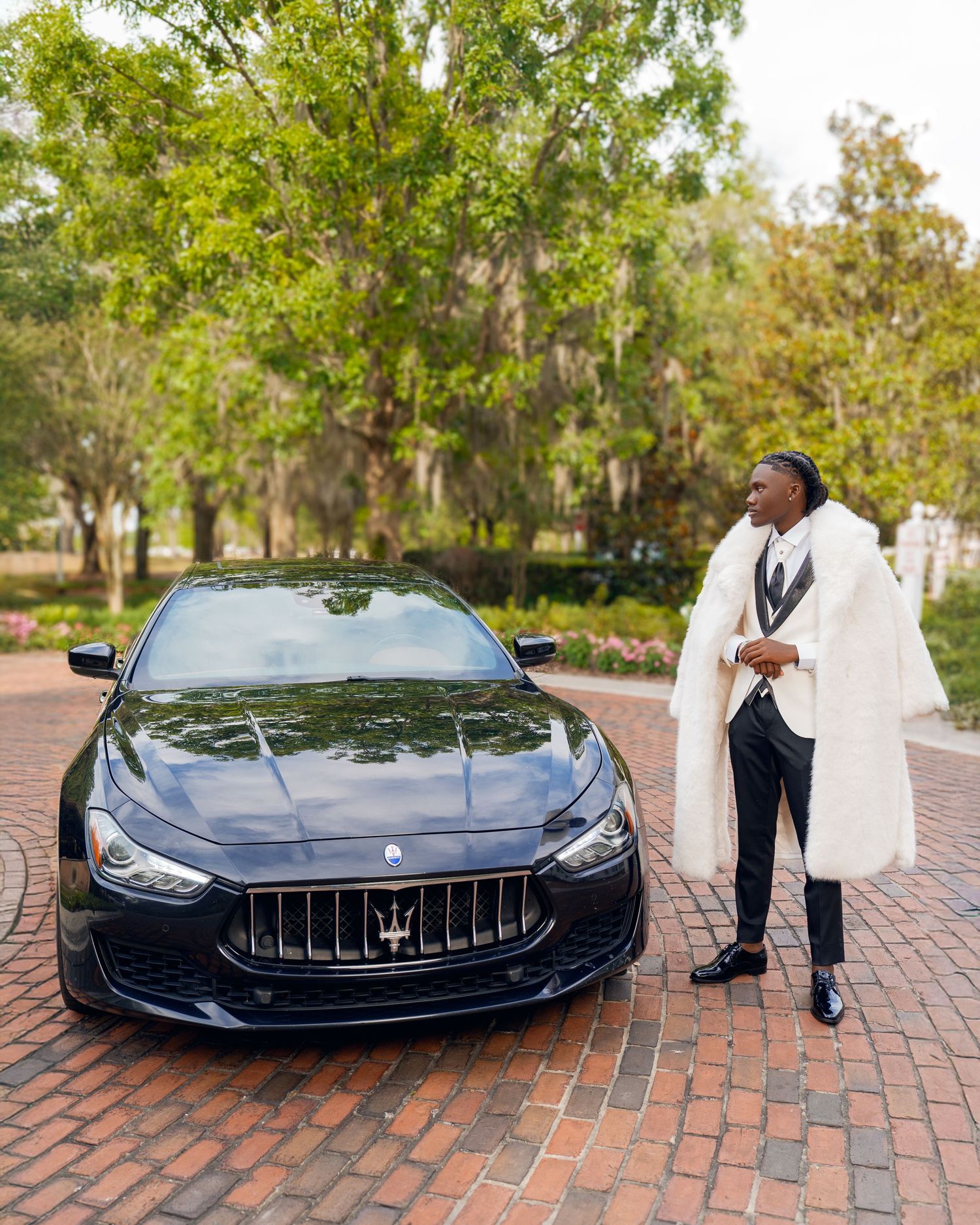 Man in a white fur coat and black suit stands beside a black Maserati on a brick driveway under leafy trees.
