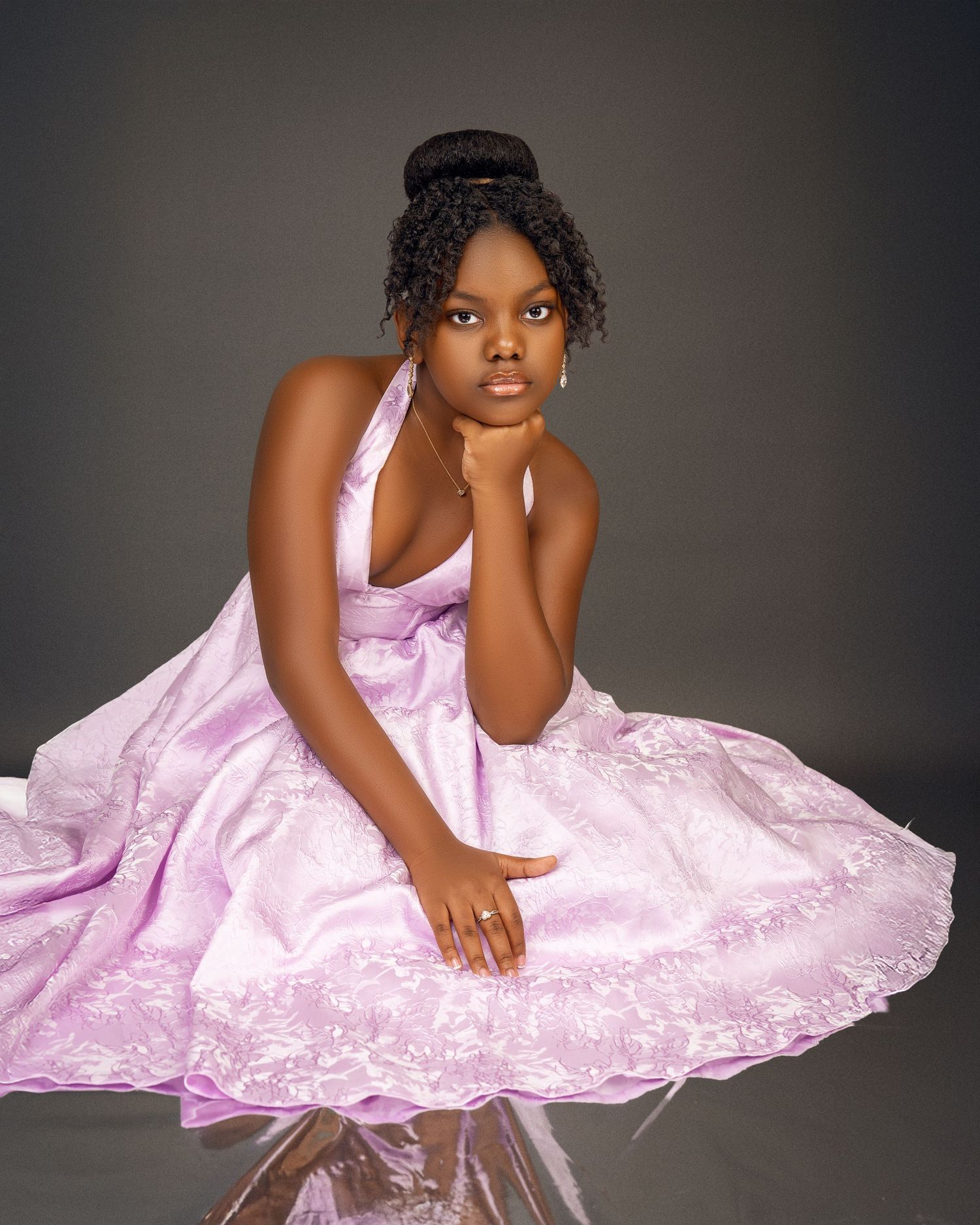 Young girl in a lilac lace halter dress sits with her chin on her hand in a graceful studio portrait.