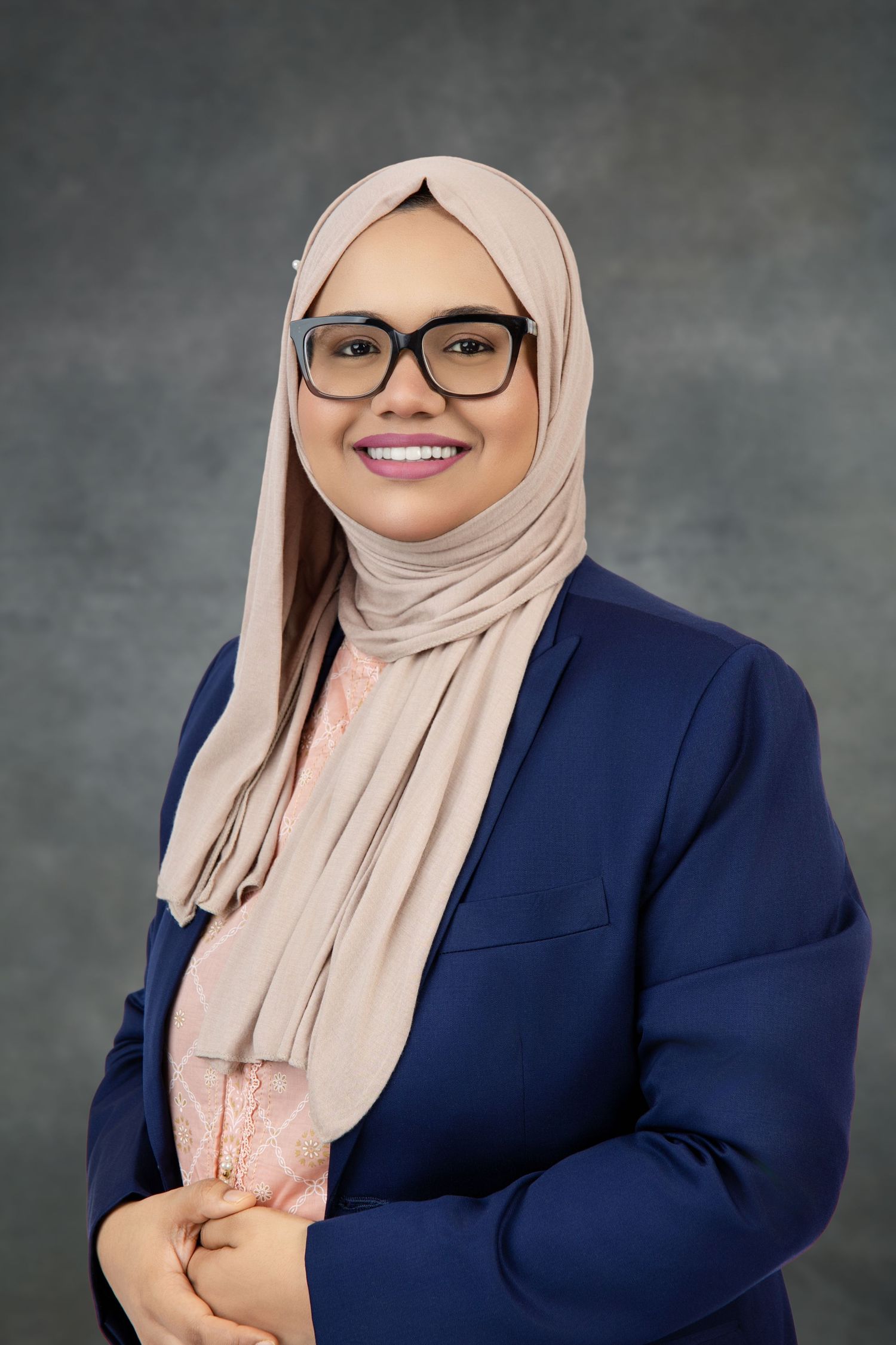 Smiling woman in a beige hijab, glasses and navy blazer poses with clasped hands in a professional portrait.
