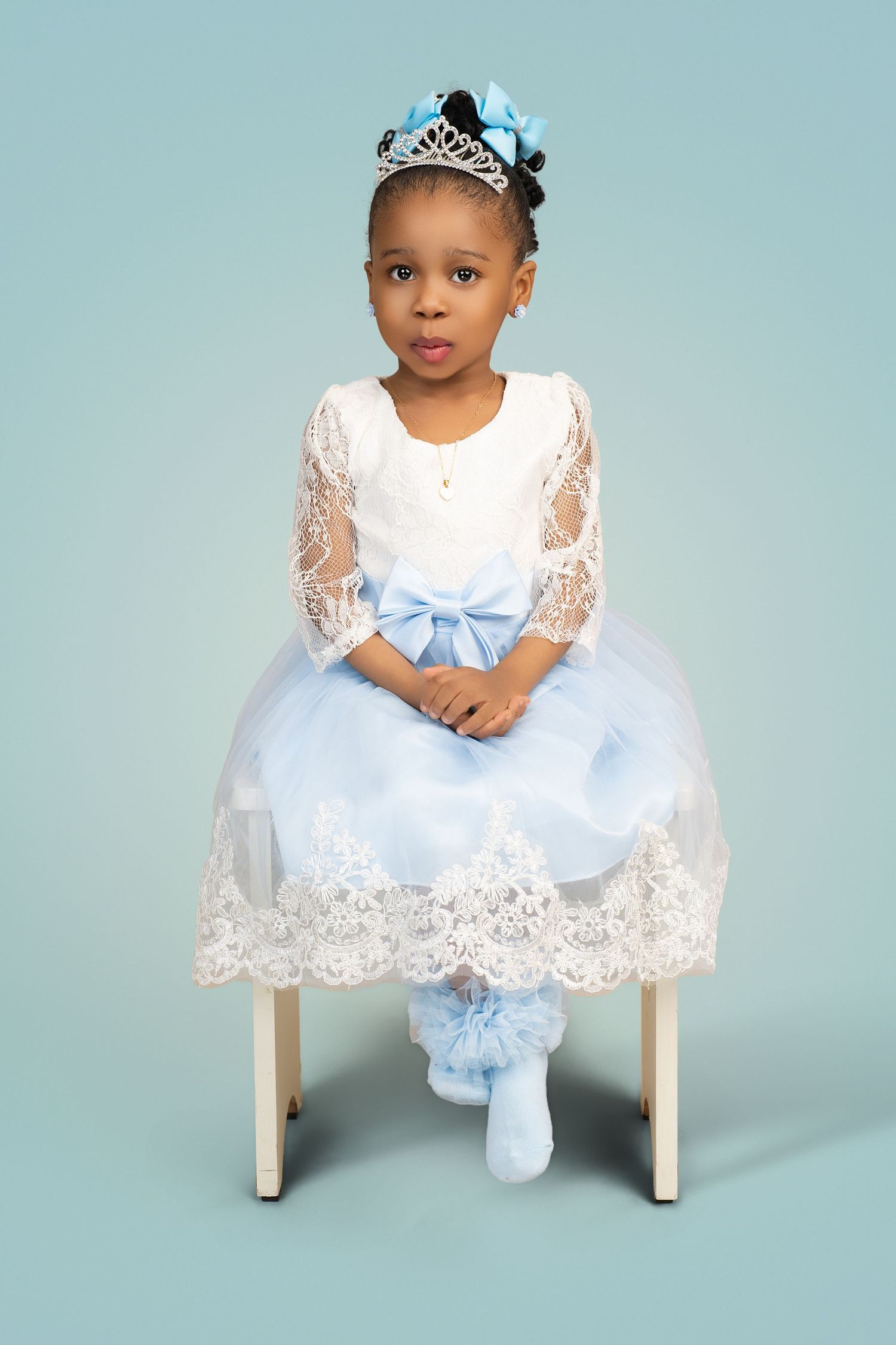 Little girl in a sparkling tiara and blue bow, wearing a white lace dress with a light blue tulle skirt, seated on a white stool.