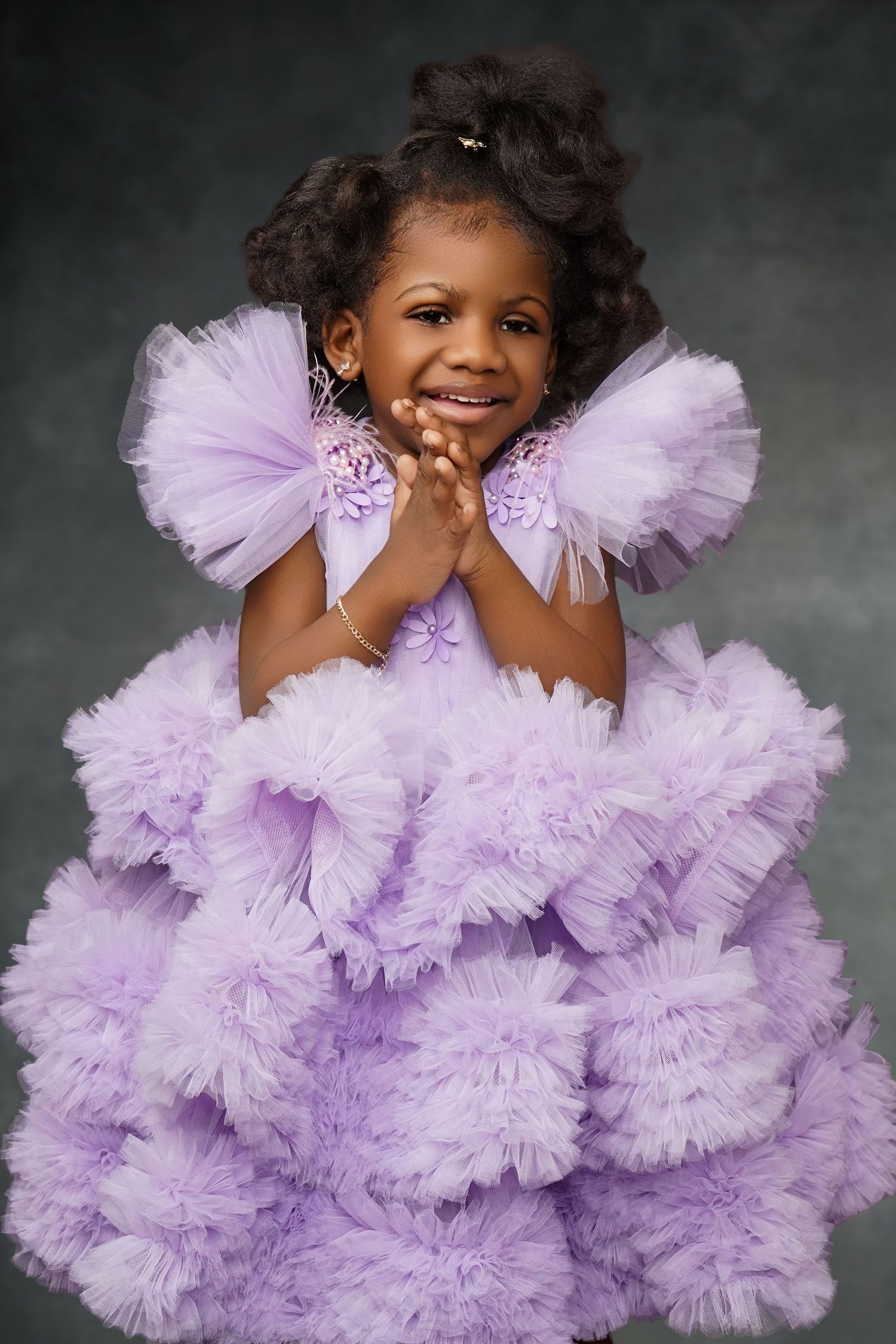 Smiling little girl with curly hair clasps her hands in a lavender tulle ruffle dress against a moody textured backdrop.