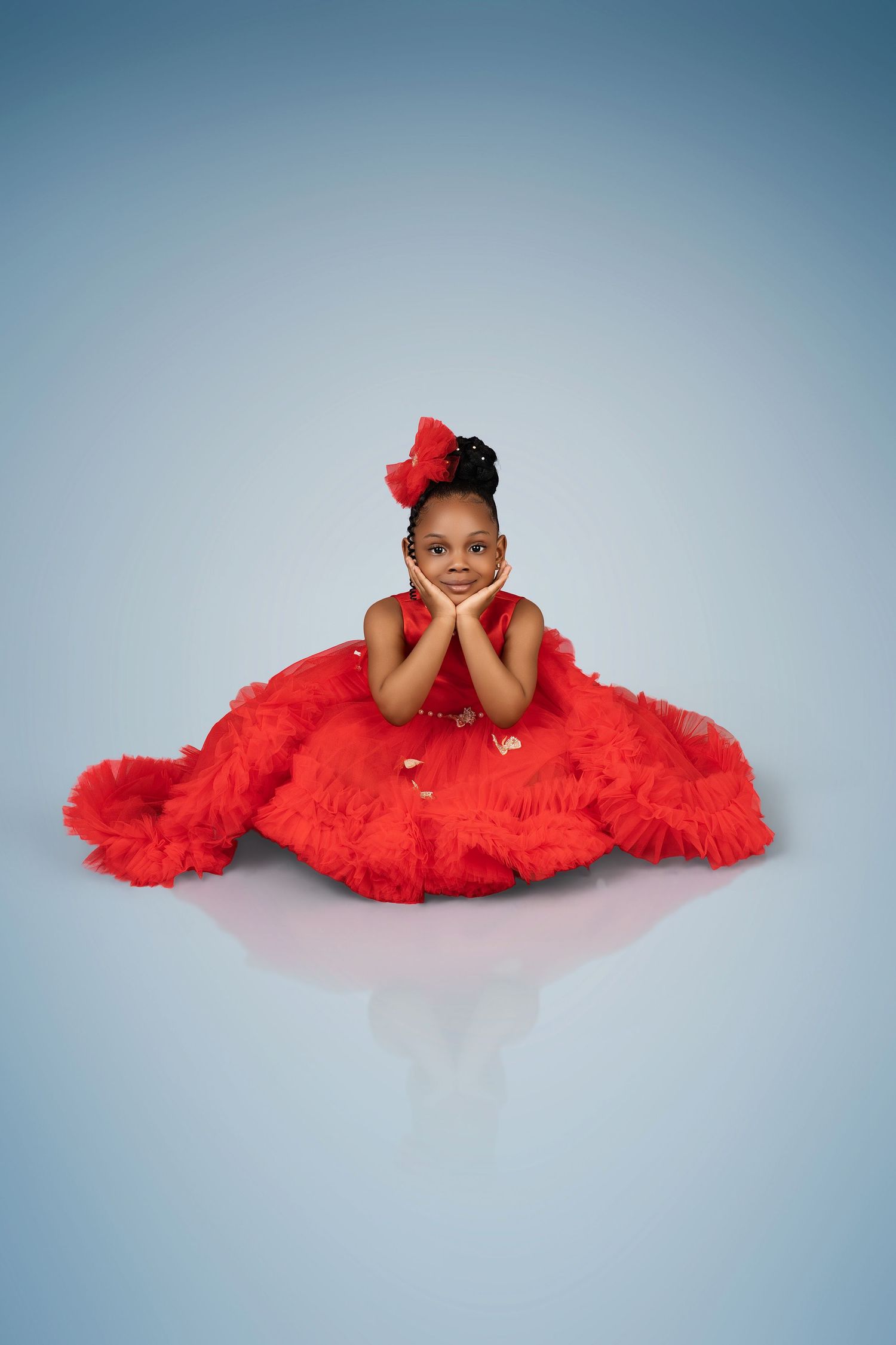 Smiling little girl in a ruffled red tulle gown and red hair bow rests her chin on her hands in a studio portrait.