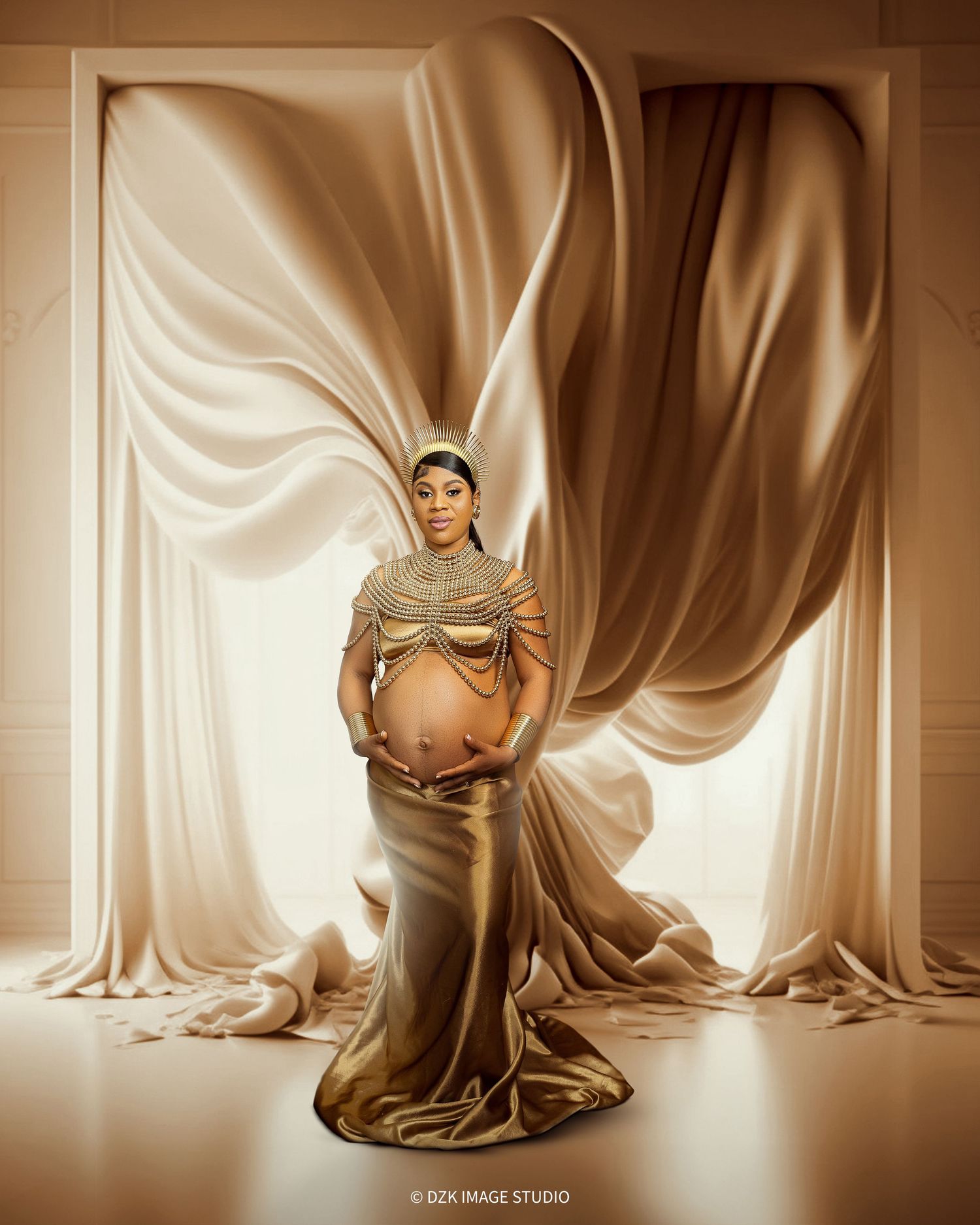 Pregnant woman in a gold beaded top and metallic gold skirt holds her belly before flowing champagne silk drapes in a studio.